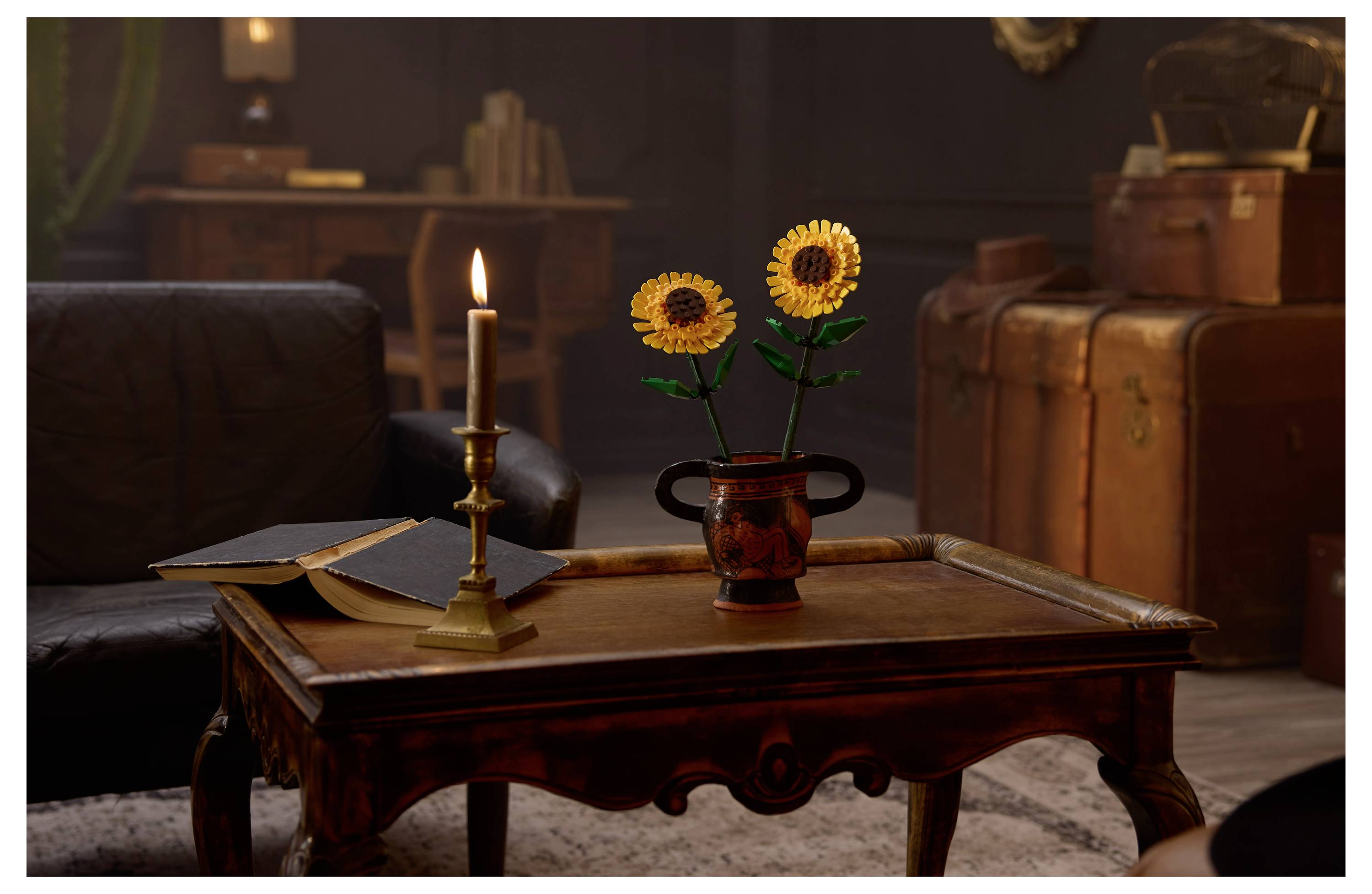 A dimly lit room with a wooden table, a candle, an open book, and a vase with sunflowers. Stacks of luggage and books are visible in the background.