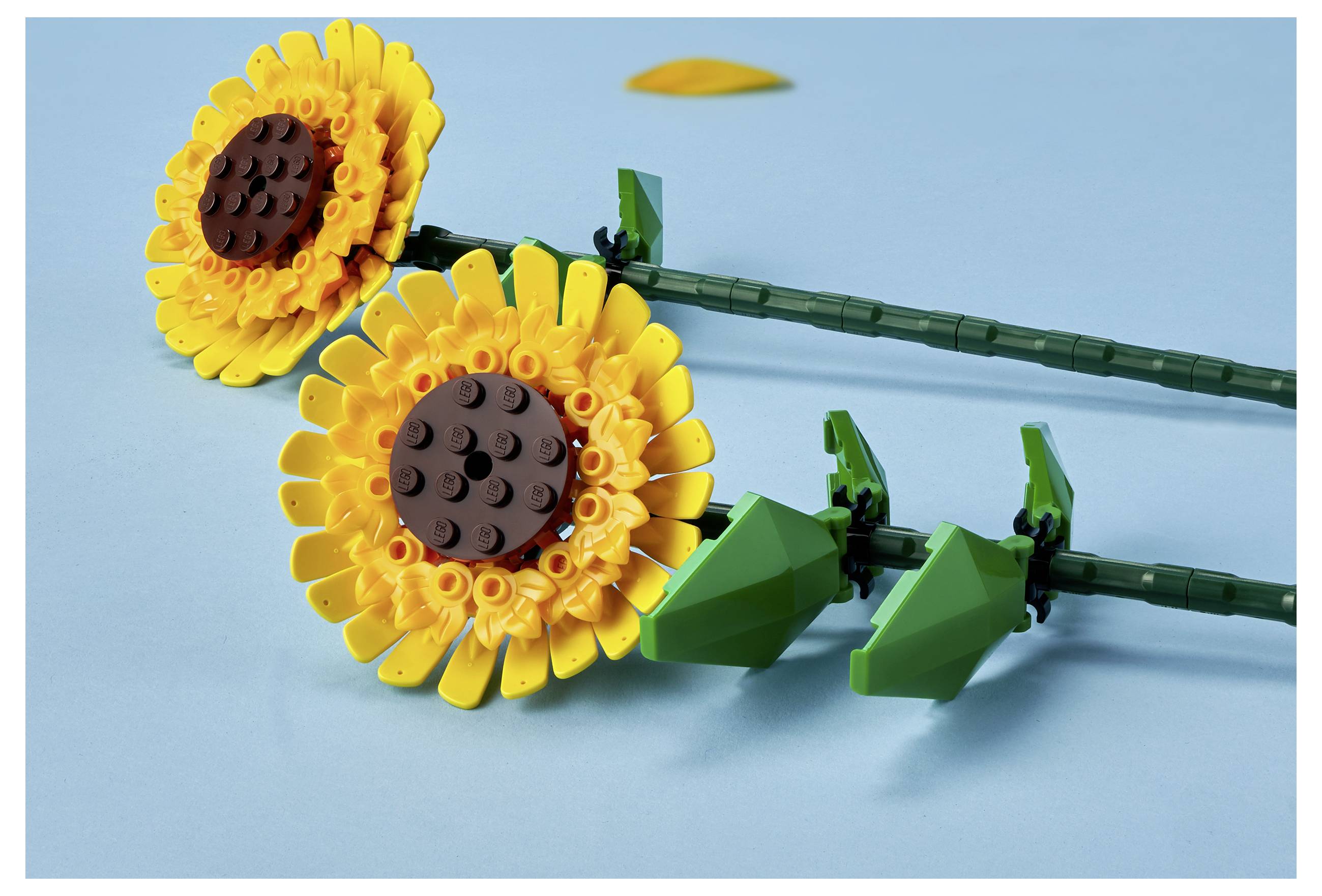 Two LEGO sunflowers with yellow petals and brown centers lie on a light blue background, showcasing detailed stem and leaf designs.