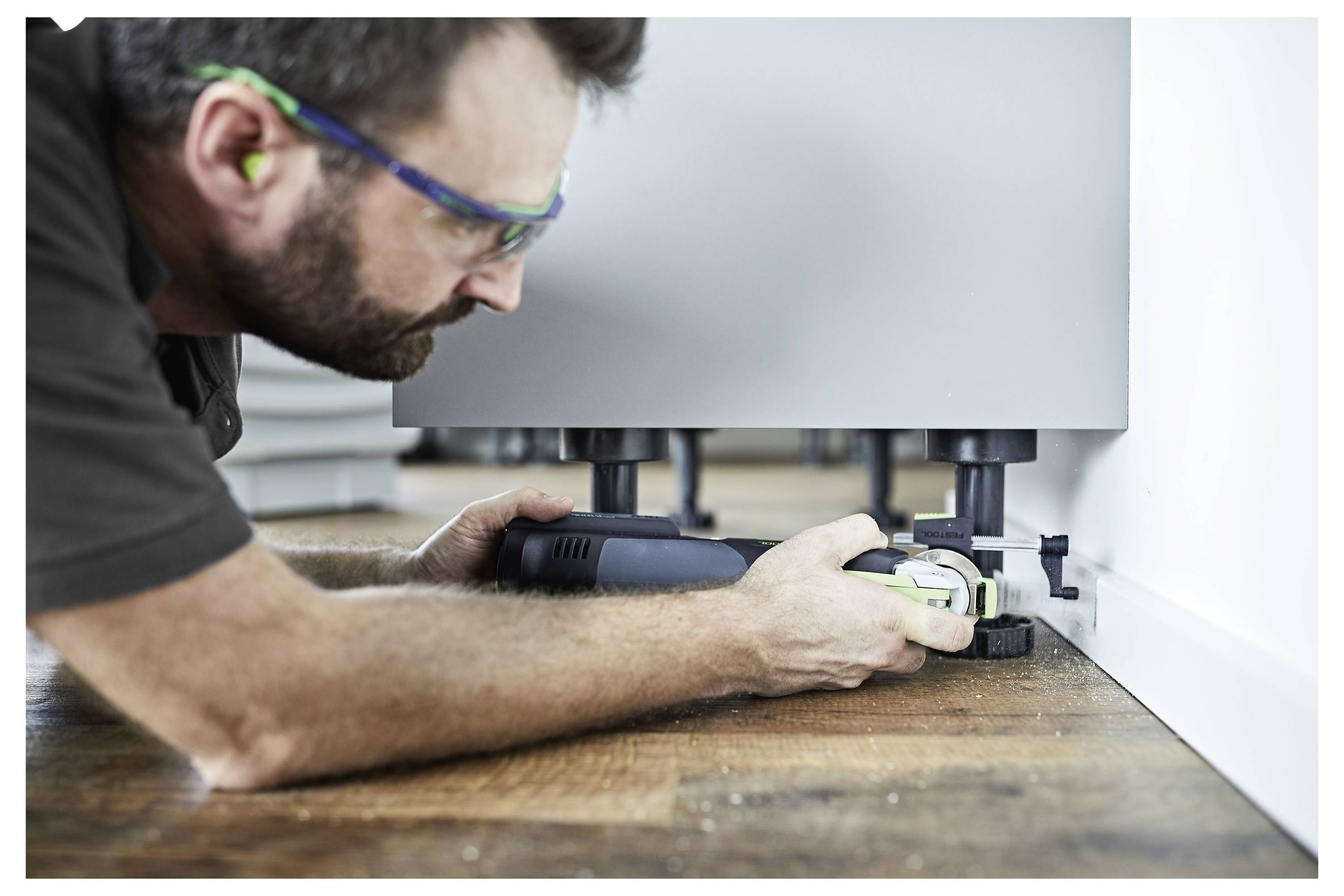 Man in protective glasses using a power tool to adjust furniture legs on a wooden floor in a room.