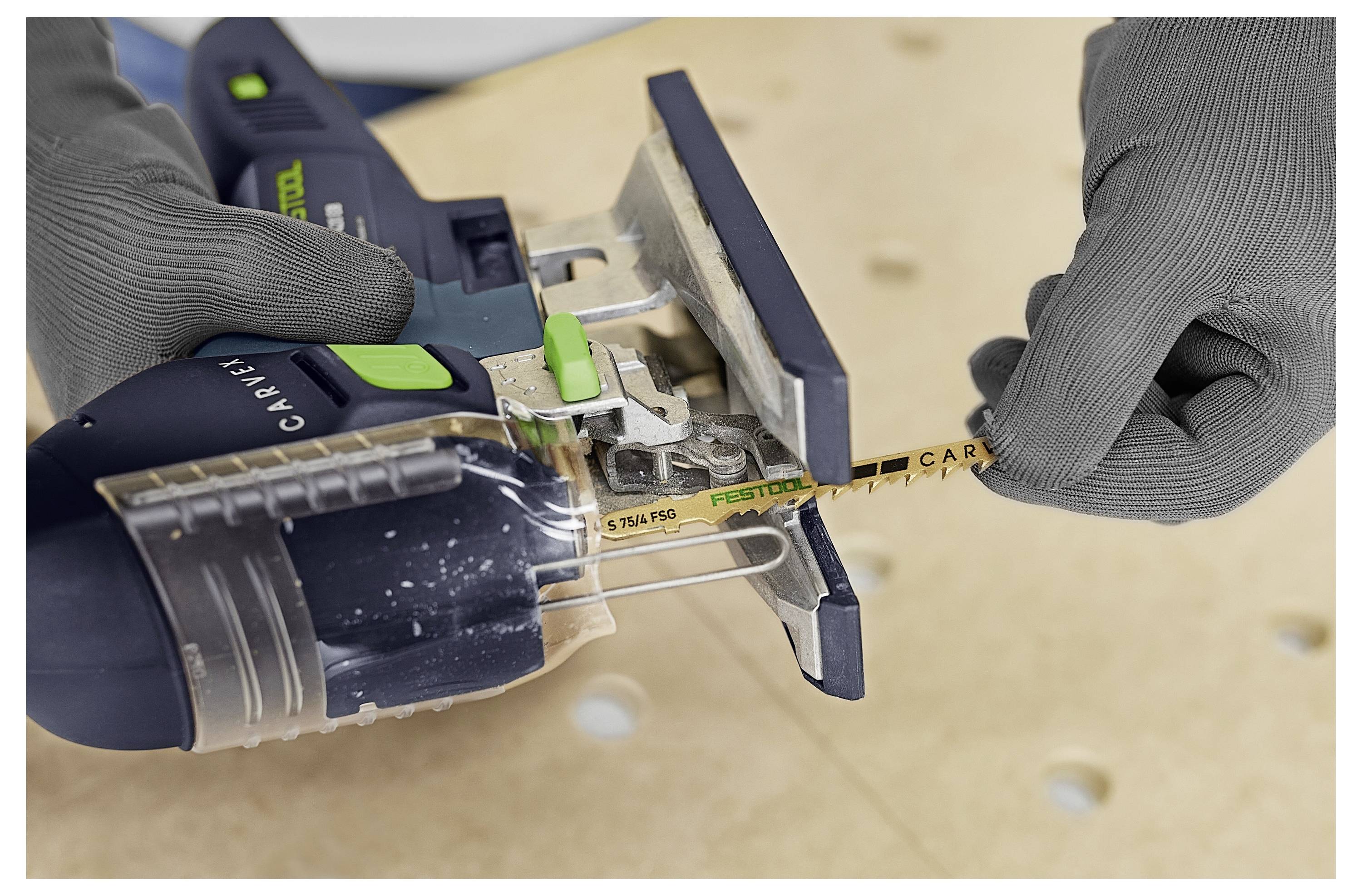 A person in gray gloves inserts a Festool blade into a jigsaw on a wooden surface, suggesting a focus on tool setup or maintenance.