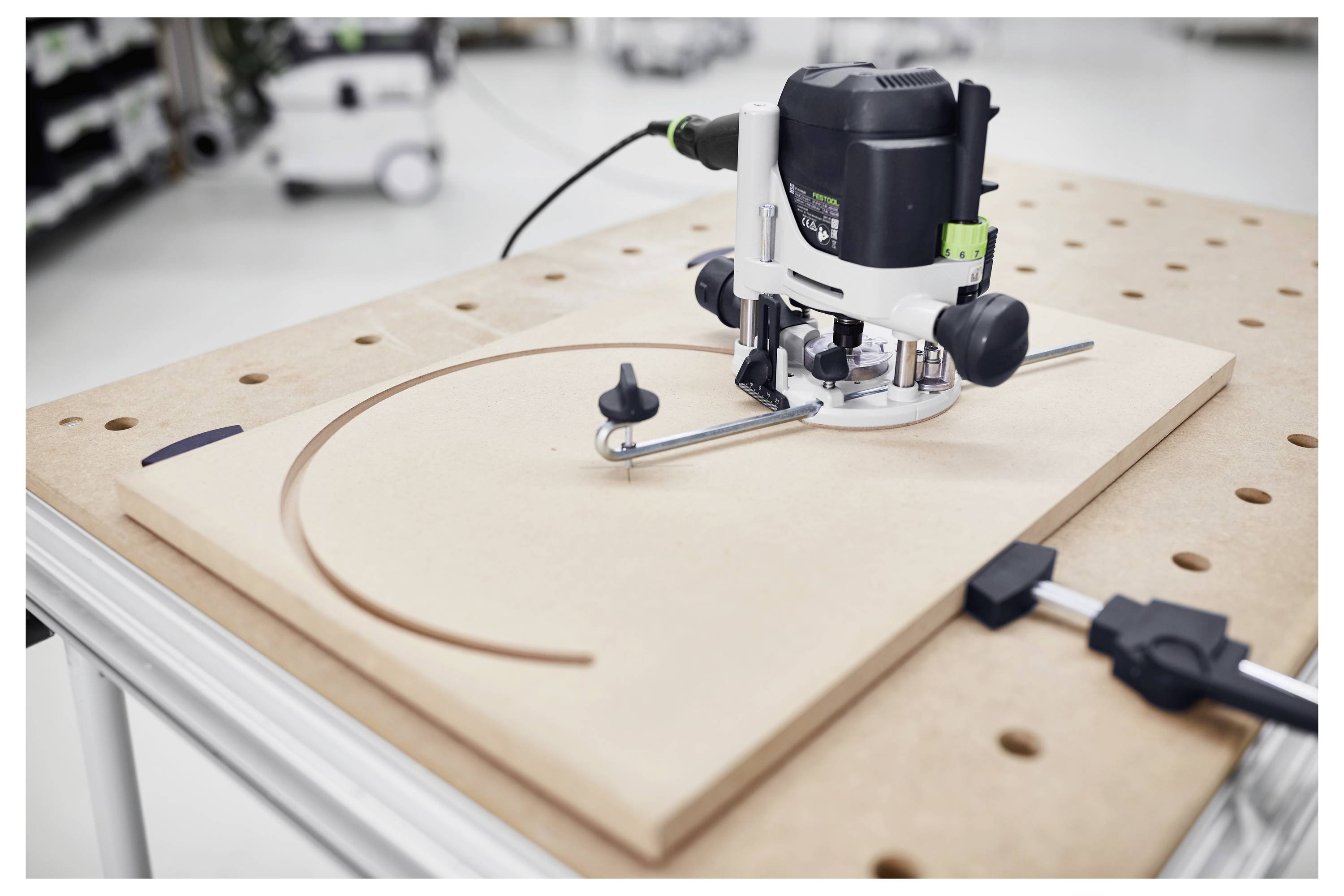 A router on a workbench is cutting a curved groove into a wooden board, controlled by a metal guide for precision.