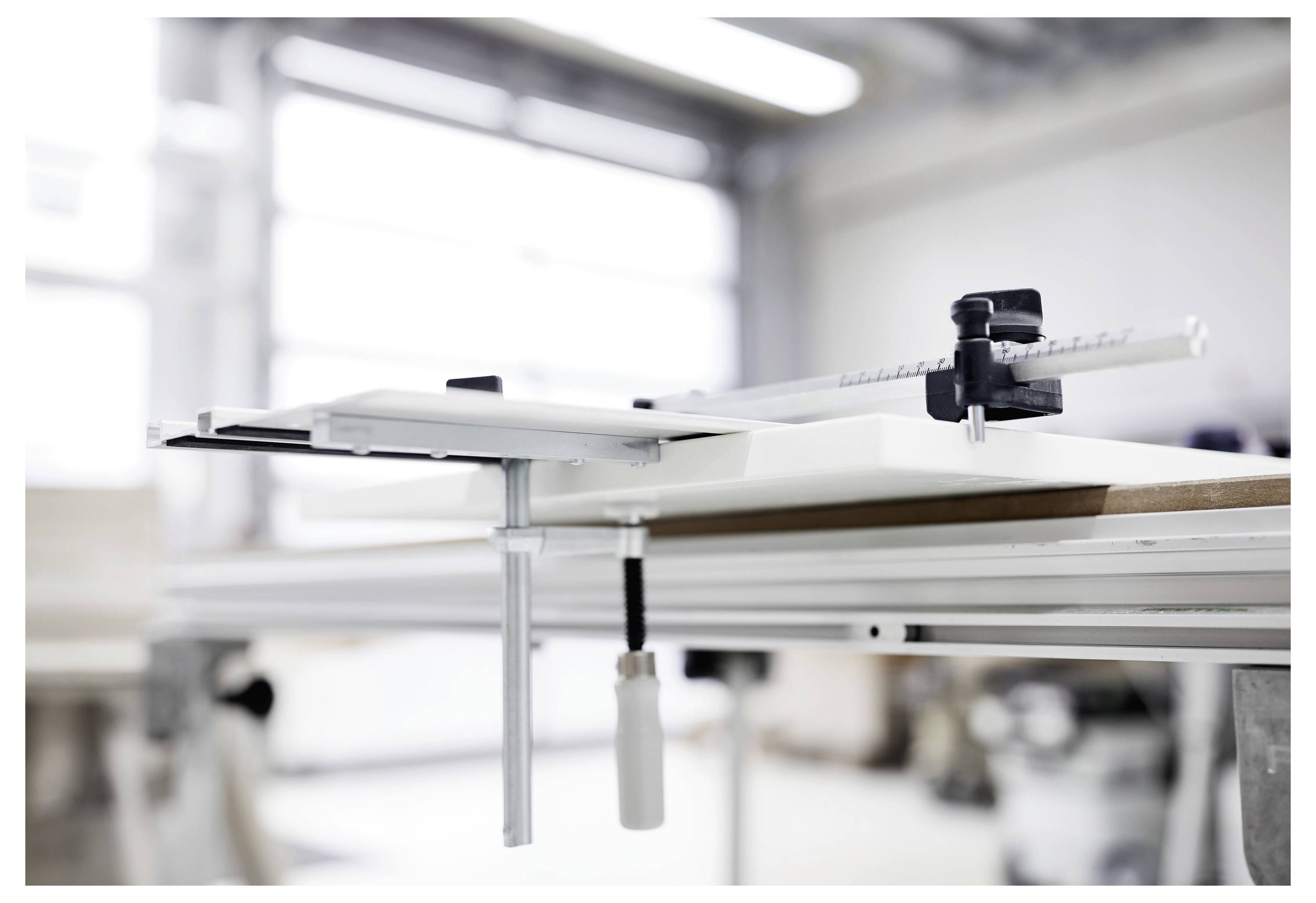 Close-up of a table saw guide with measurement tool, clamped to a workbench in a bright workshop setting.