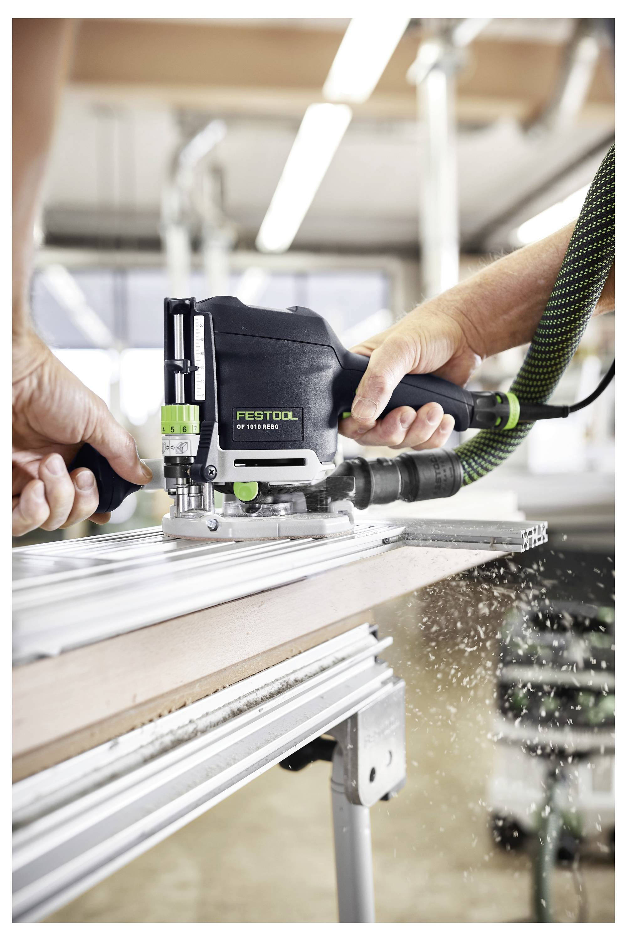 A person uses a Festool router to cut a wooden board. The tool is positioned on a metal guide for precision, in a workshop setting.