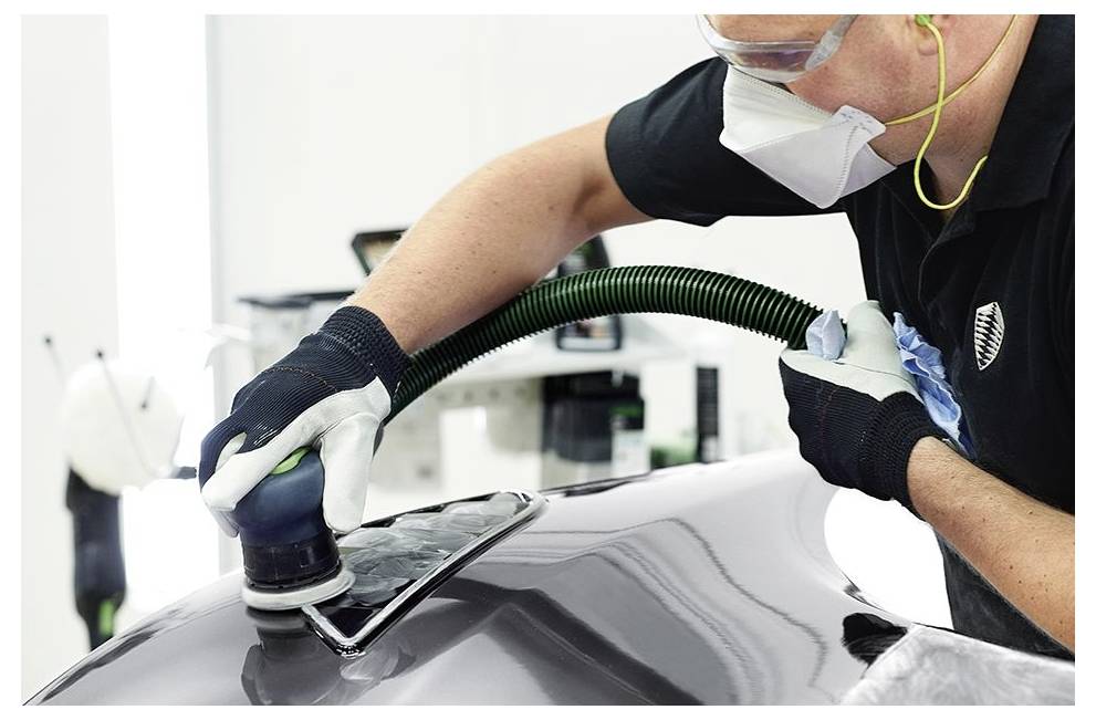 A person in protective gear polishes the surface of a car hood with an electric buffer in a workshop setting.