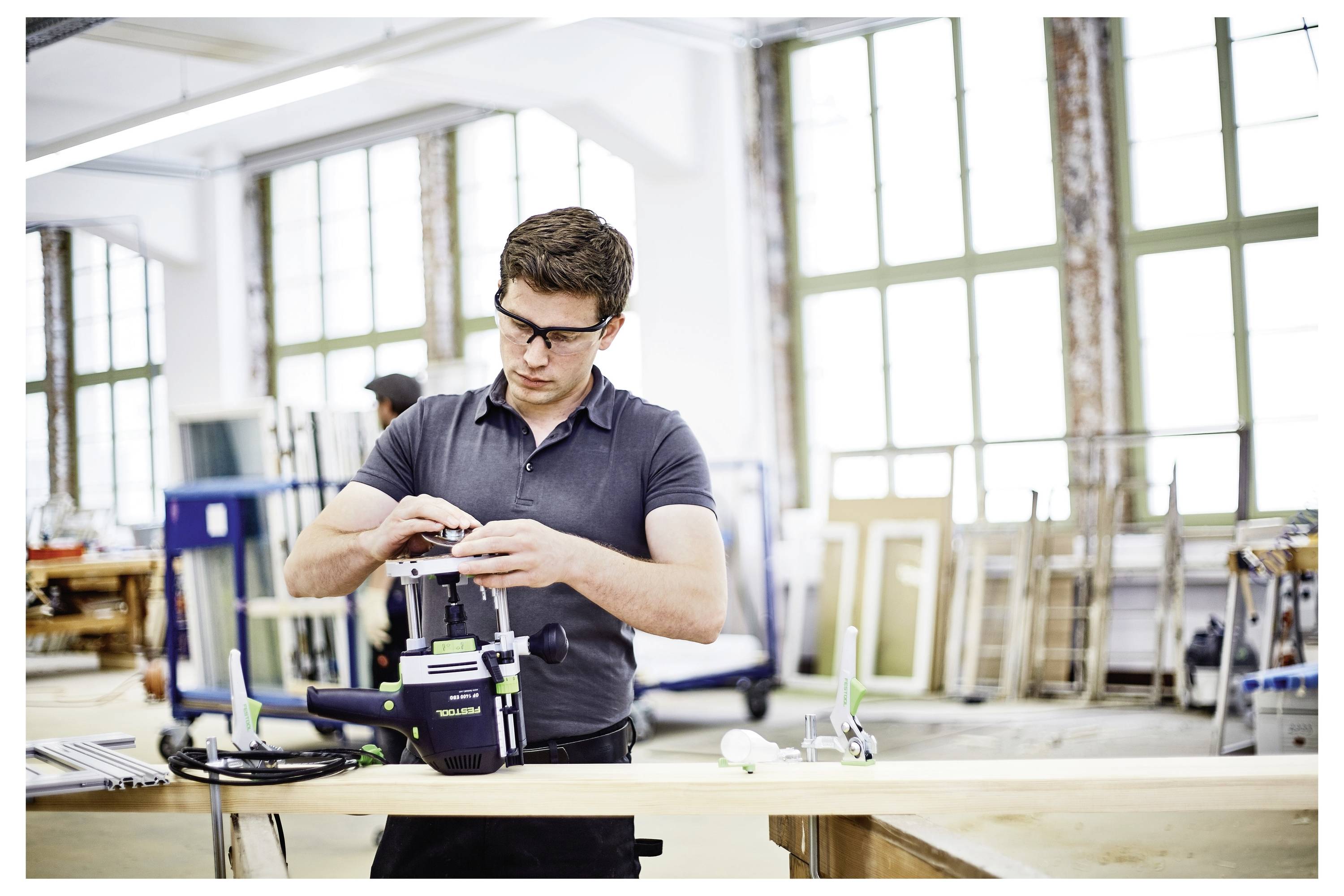 A person wearing safety glasses assembles a tool on a workbench in a workshop setting with large windows and various equipment around.