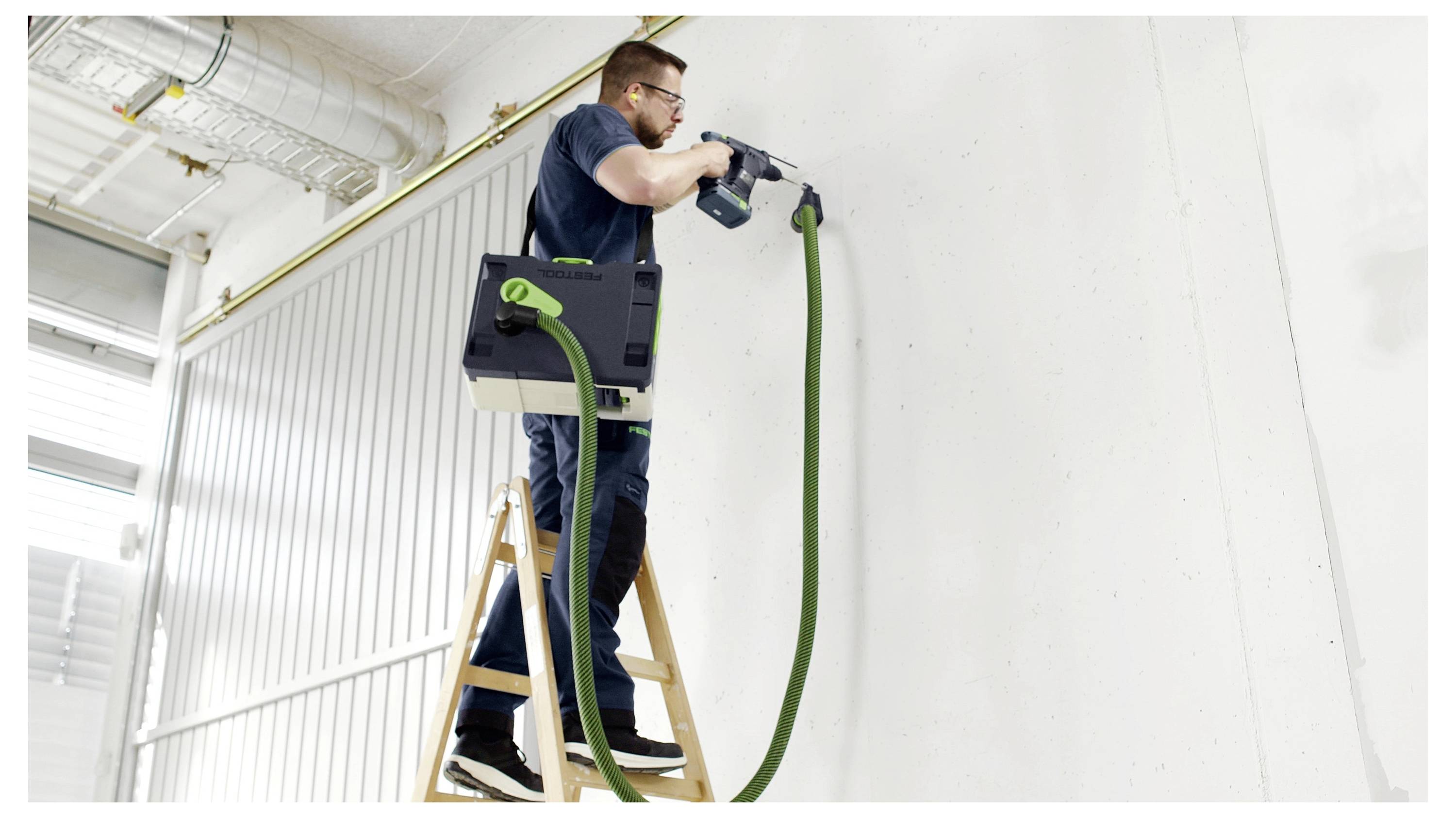 A man standing on a ladder uses a drill on a wall, with a vacuum system attached to collect dust, inside a bright industrial space.