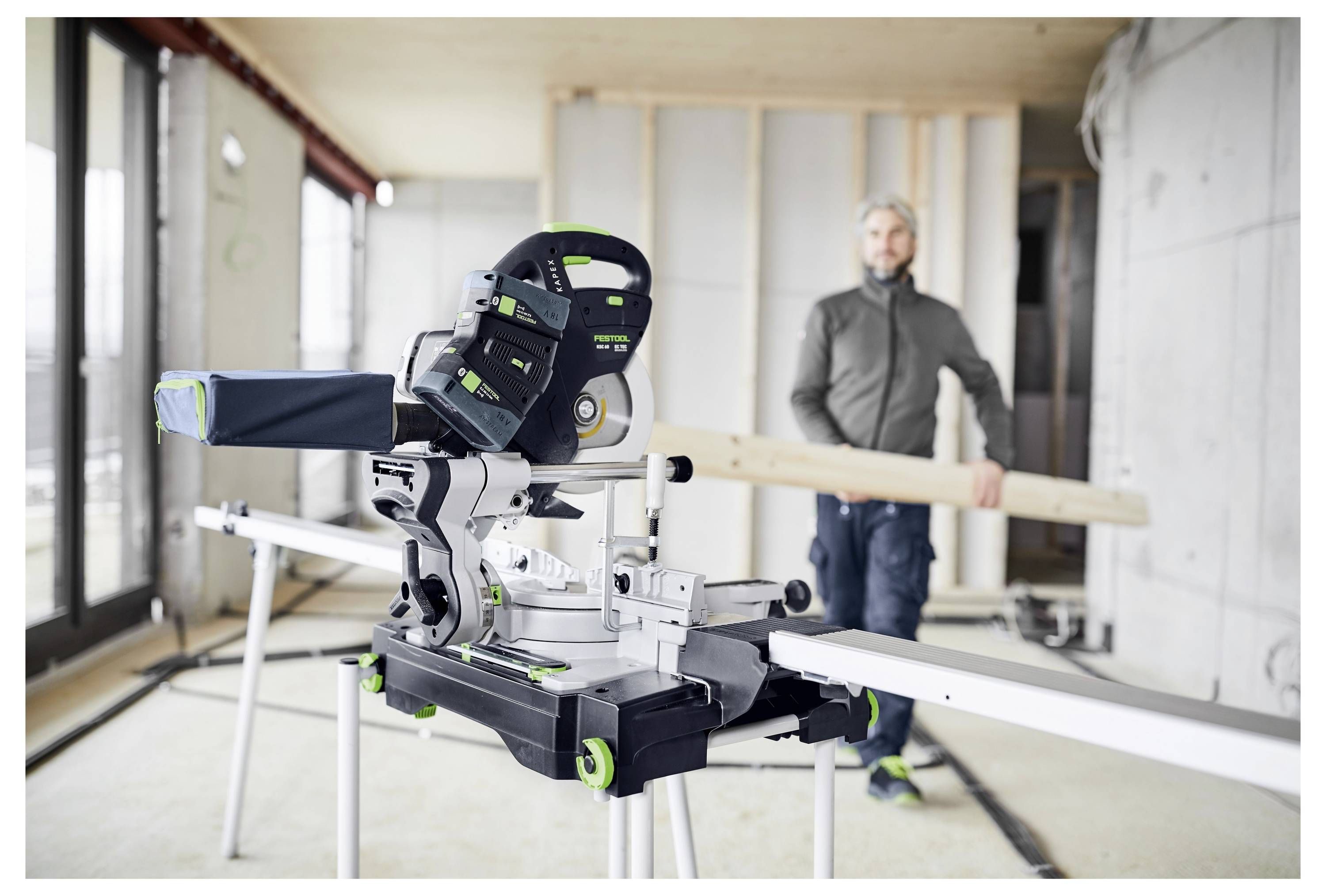 A person in a partially constructed room carrying a wooden beam. In the foreground, a miter saw is positioned on a workstation.