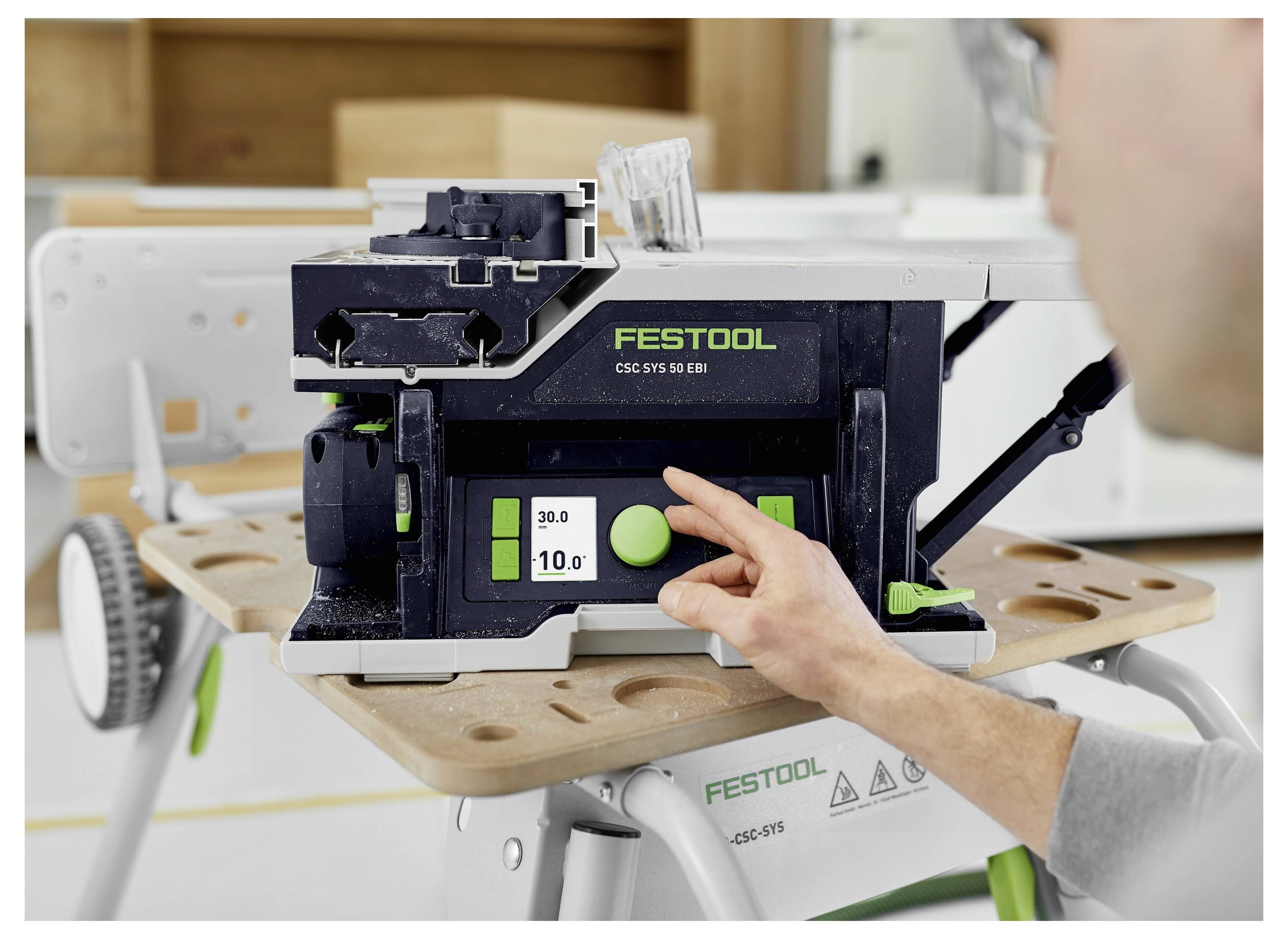 A person is adjusting a Festool CSC SYS 50 EB cordless table saw, focusing on the digital display showing depth settings of '20.0' and '10.0'.