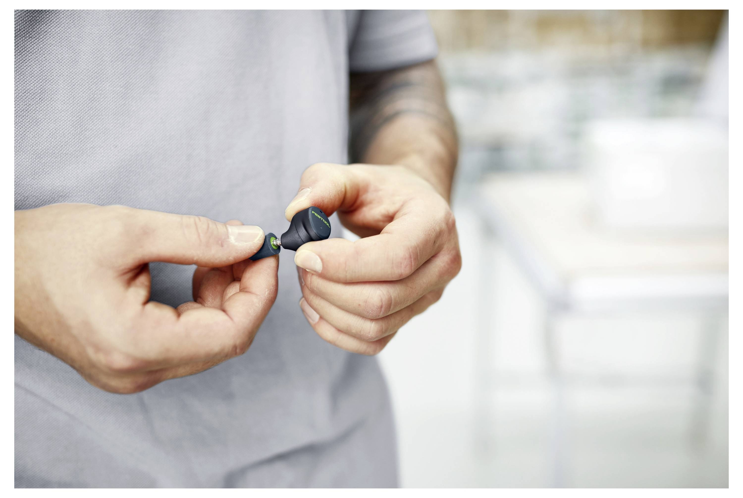 A person adjusts a small, dark wireless earbud with both hands. The background is blurred, suggesting an indoor setting.