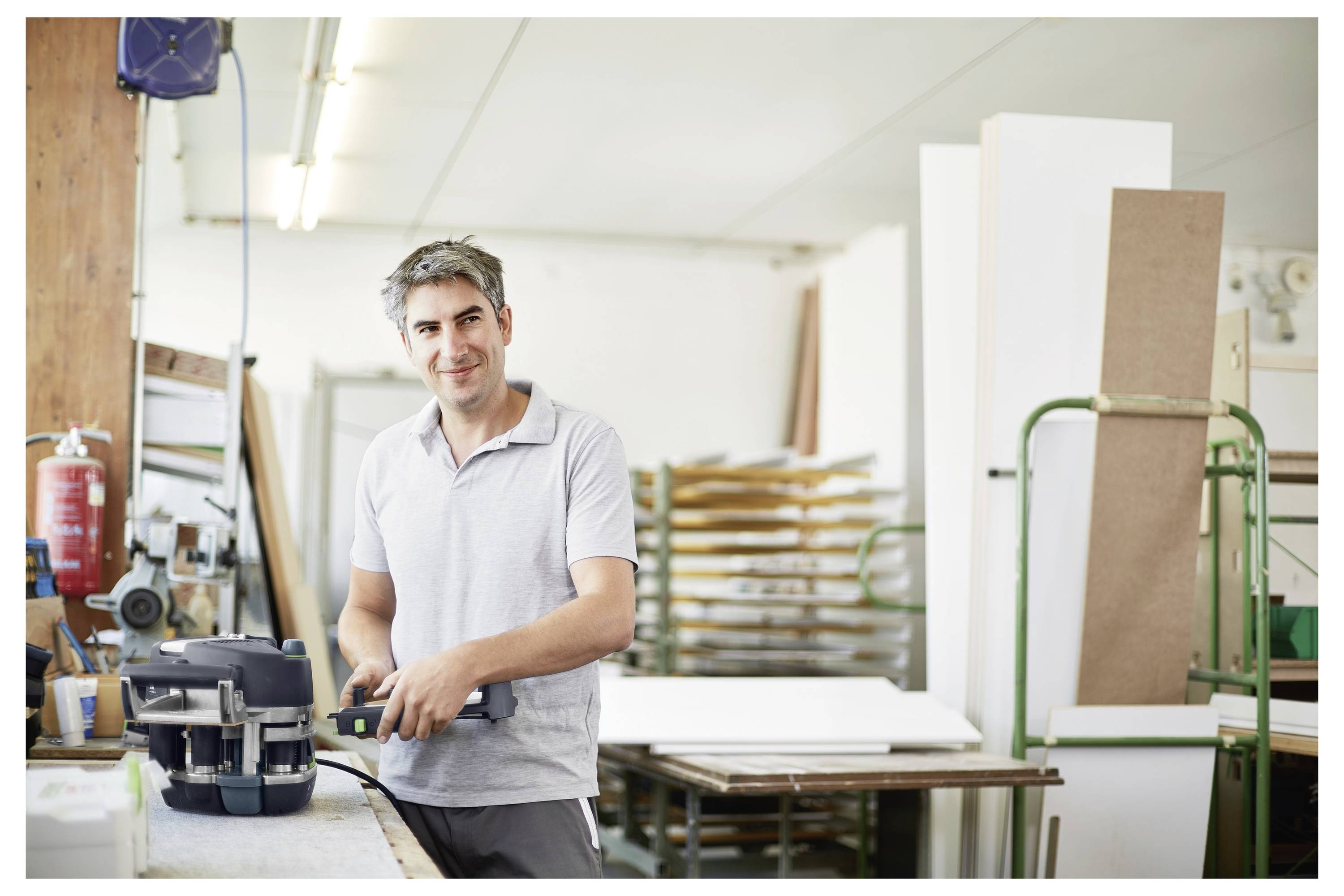 A person wearing a gray polo shirt is standing in a workshop filled with woodworking materials, holding a power tool and smiling at the camera.