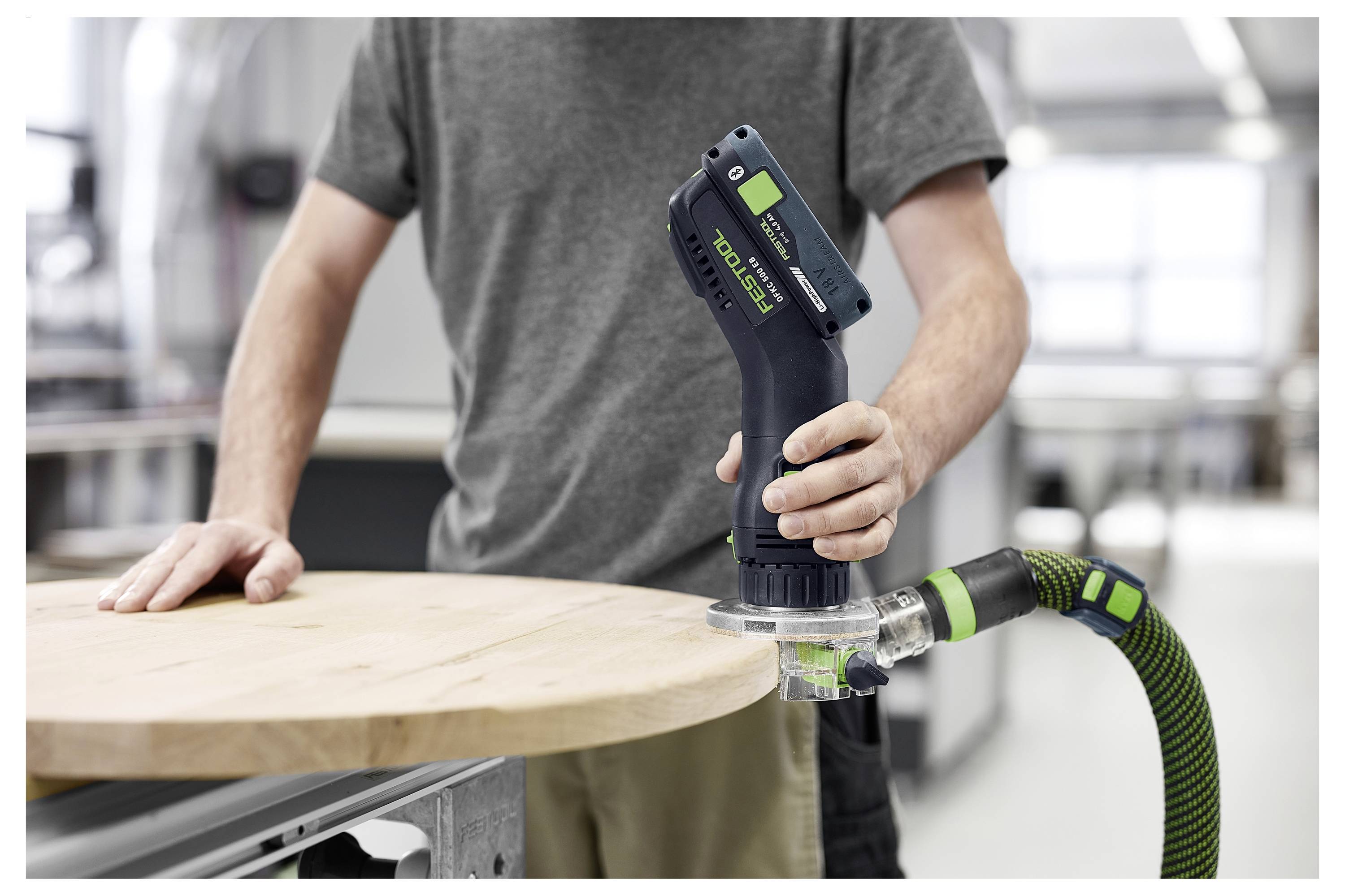 Person using a handheld sander on a round wooden surface in a workshop setting, wearing a gray shirt.
