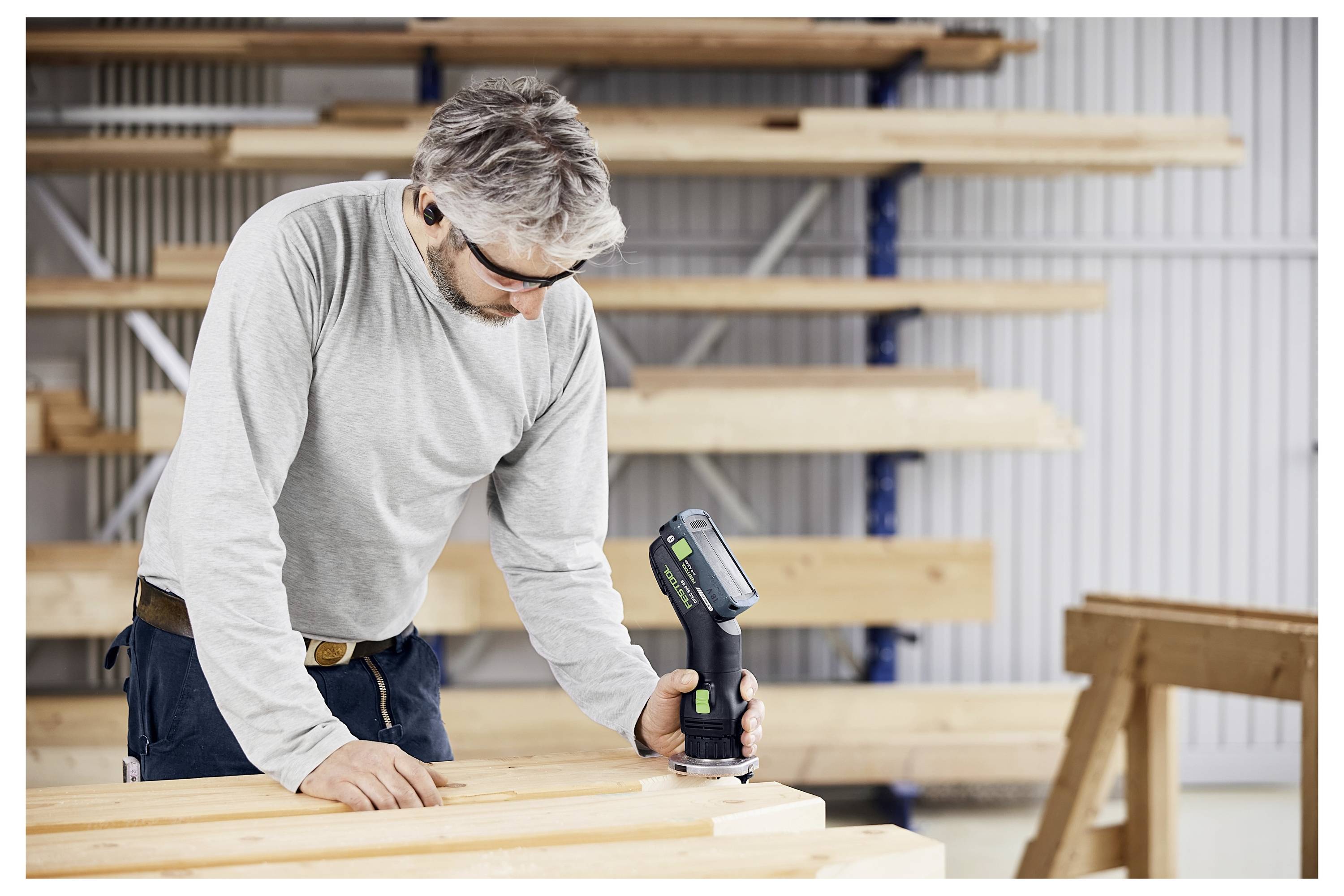 A person in safety goggles and ear protection uses a power tool on a wooden board in a workshop with stacks of lumber in the background.