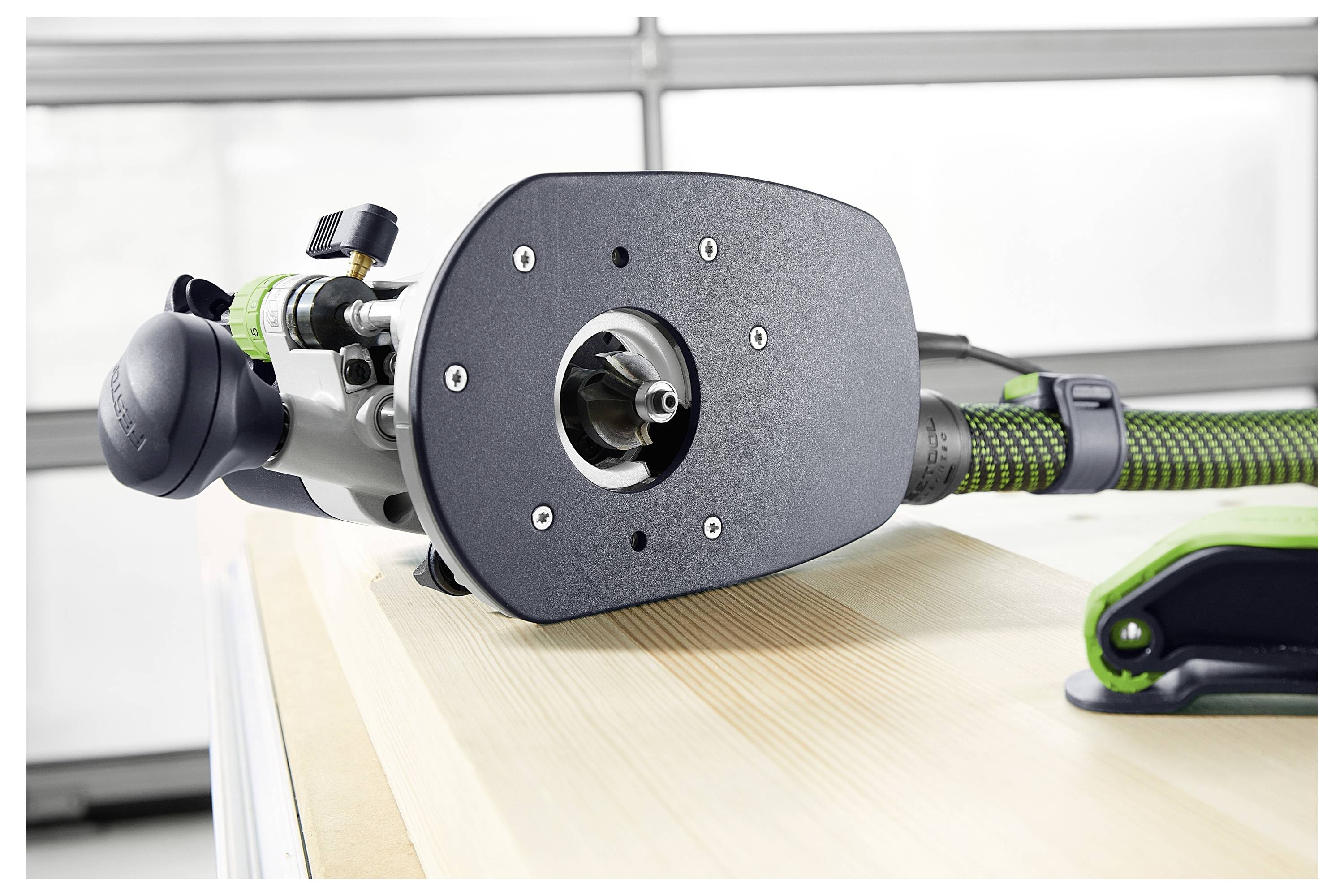 A woodworking tool, possibly a router, placed on a wooden surface, showing its base and power connection, in a workshop setting.