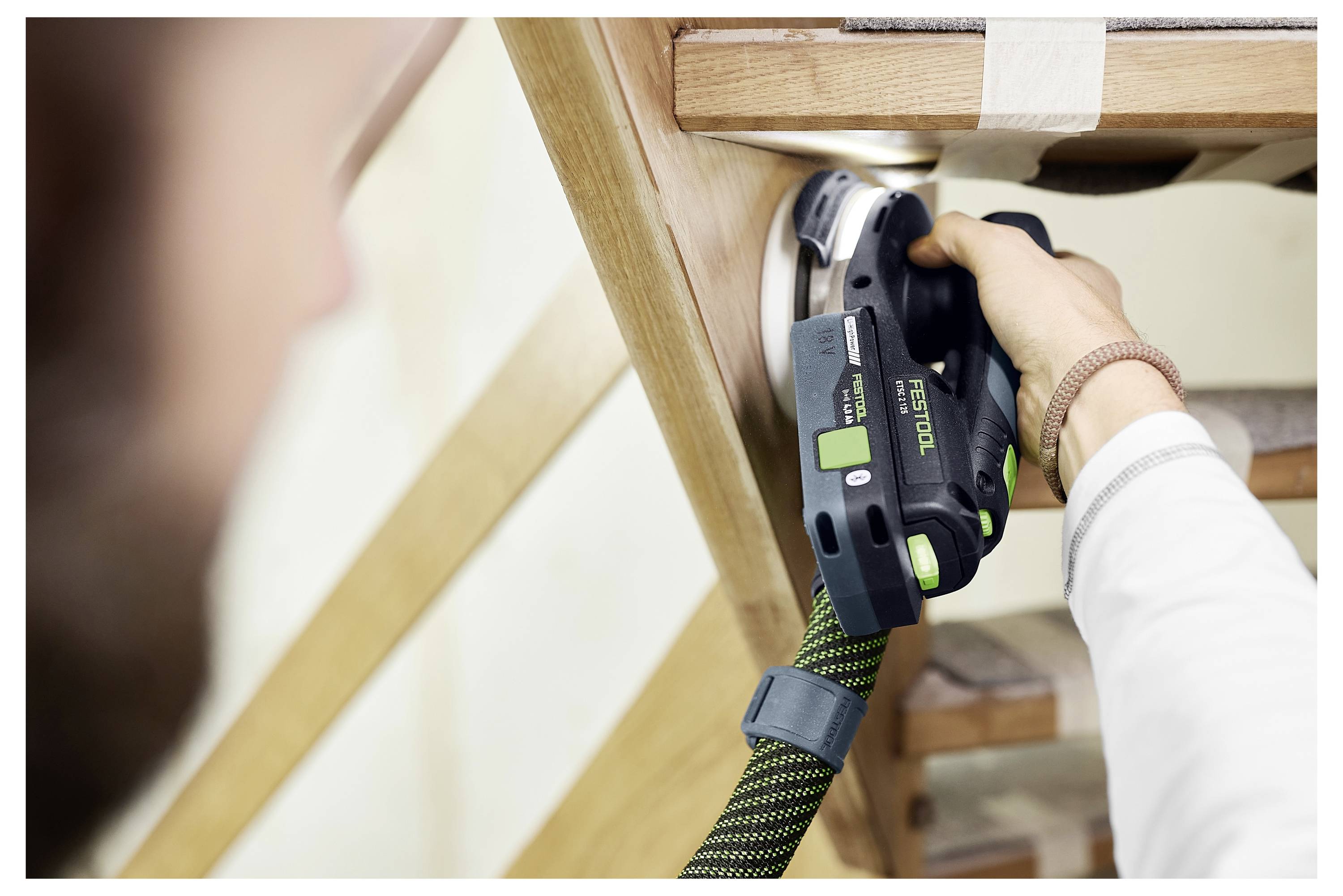 A person uses a power sander on a wooden piece, focusing on smoothing a surface. The tool is black and green, and the setting is indoors.