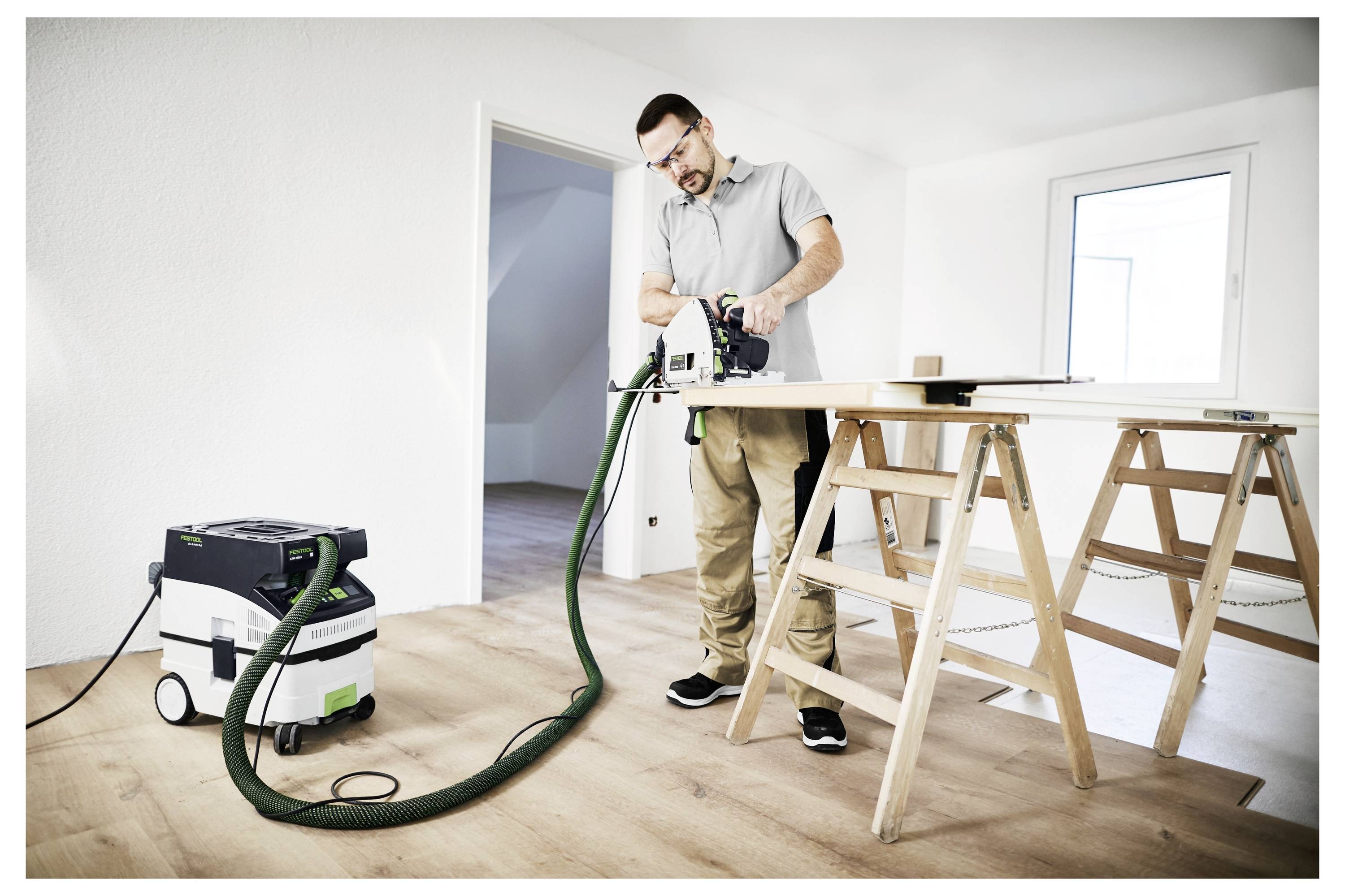 A man in safety glasses uses a power saw on a wooden board supported by sawhorses in a well-lit, white-walled room.
