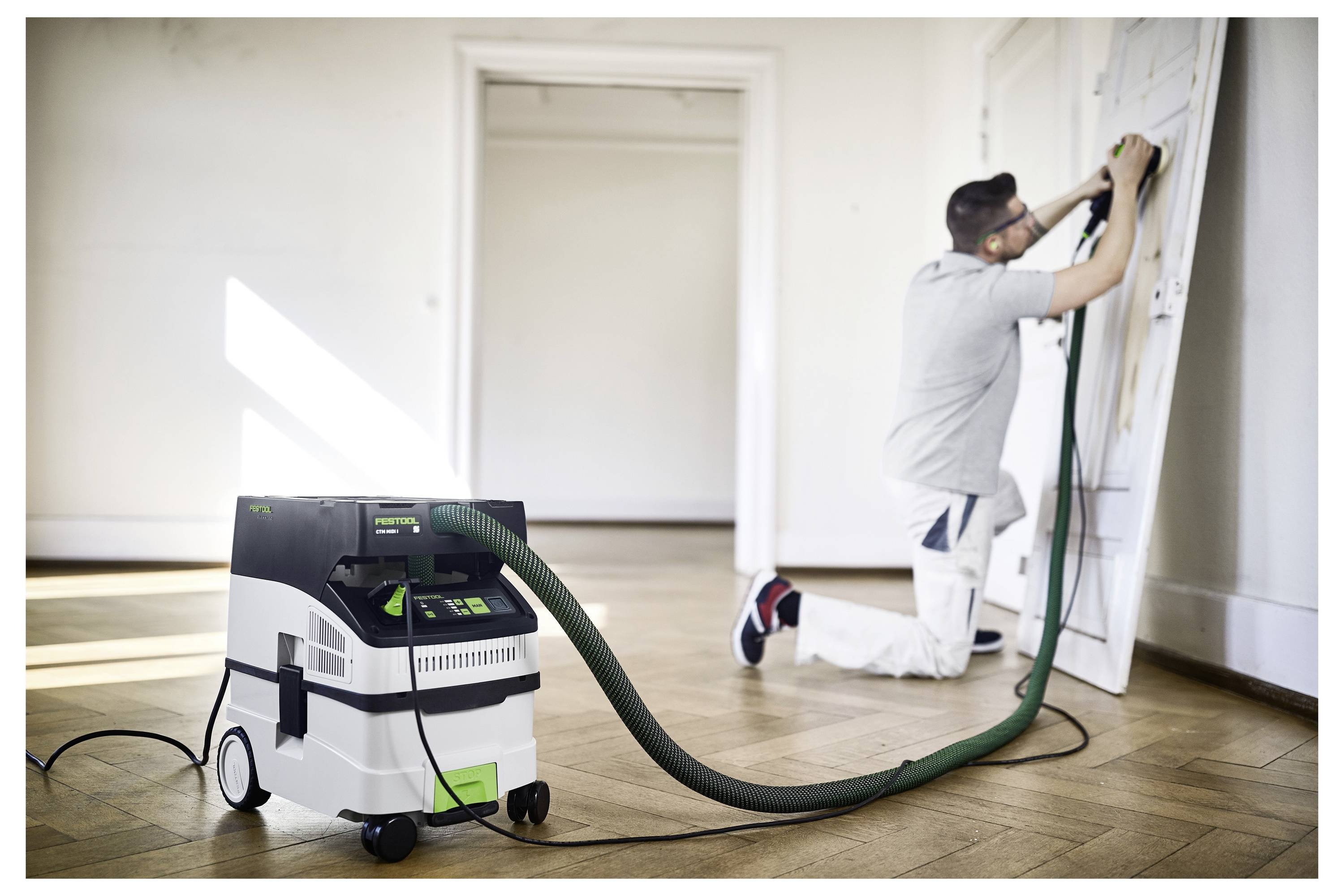 A person in work attire sands a door using a power tool, connected to a white and green dust extractor, in a bright room with wooden flooring.