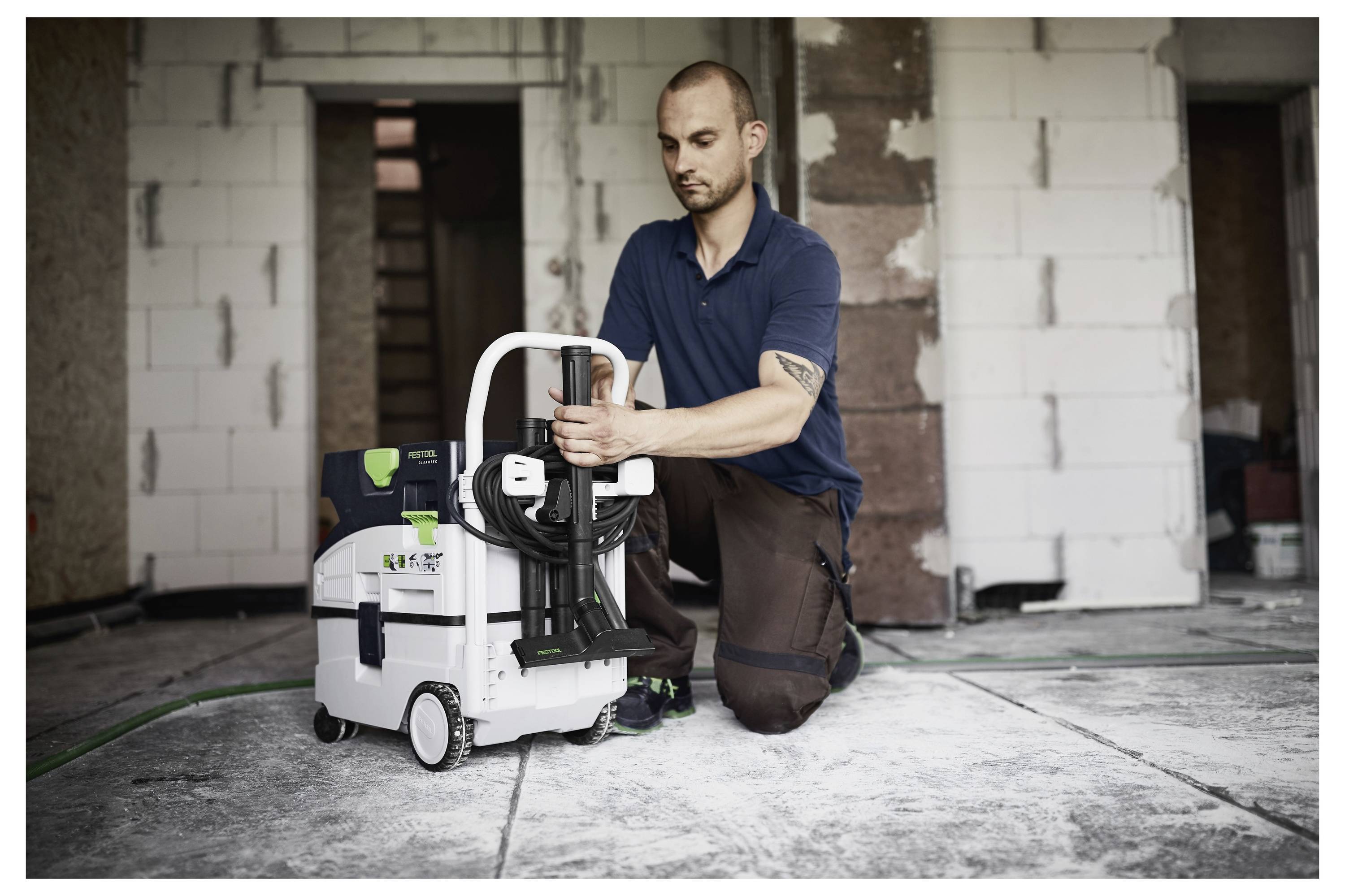 Man kneeling on a construction site floor, assembling a white portable dust extractor. Surrounds show unfinished walls and electrical cords.