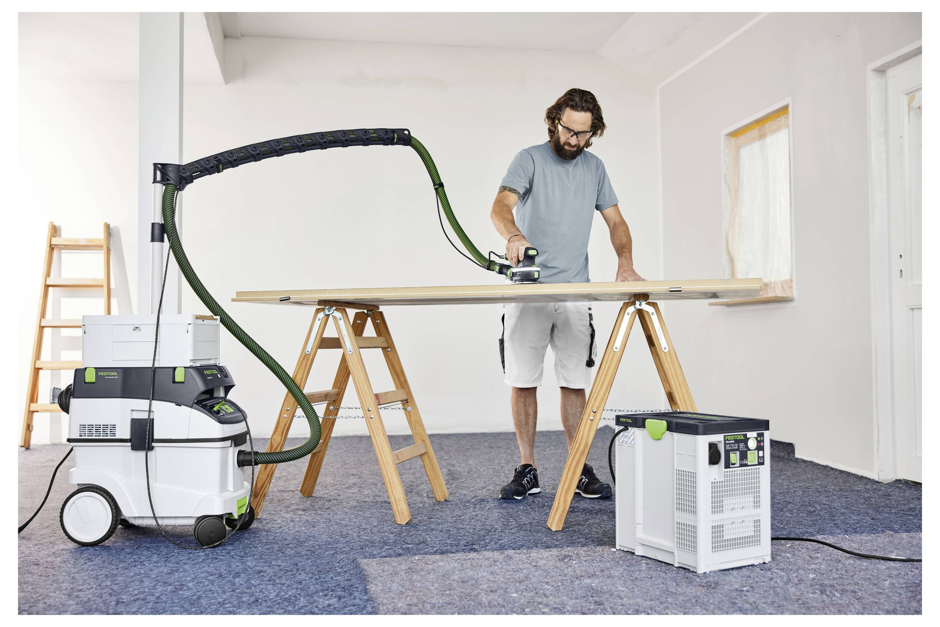 A person sanding a large wood board on sawhorses, using a sander connected to a vacuum for dust collection, in a workshop.