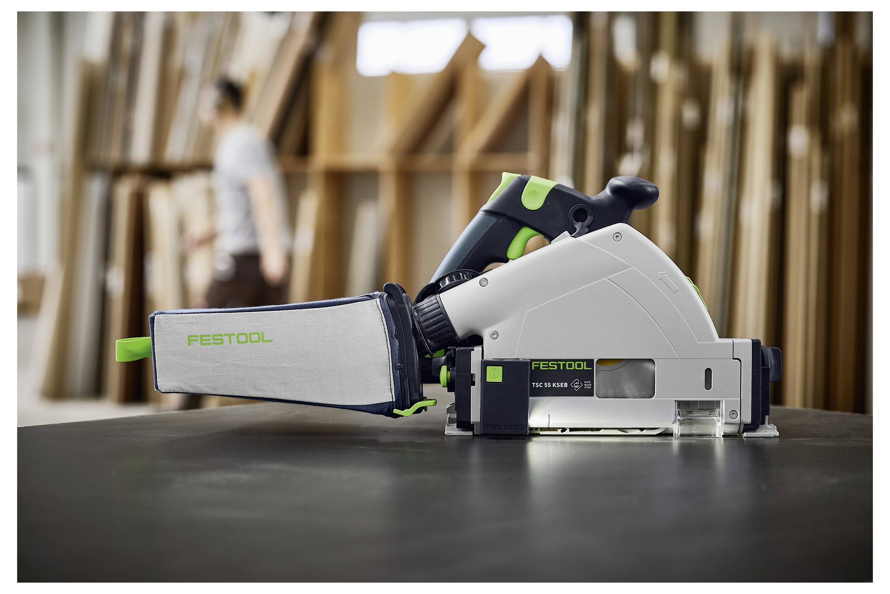 A circular saw with the brand 'Festool' on a workbench in a woodworking shop; blurred background with shelves and a person walking.