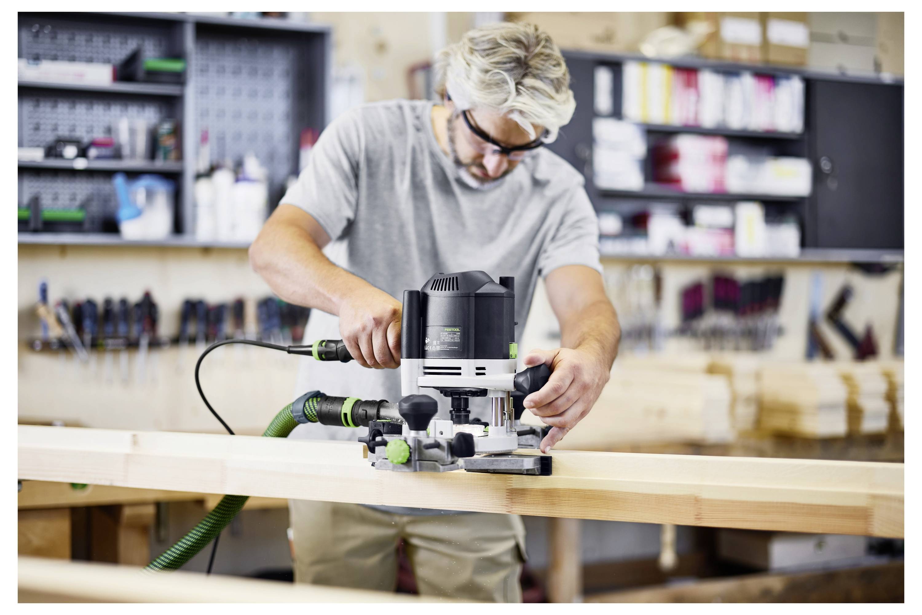 A person wearing safety goggles operates a woodworking router on a wooden plank in a workshop, surrounded by tools and equipment.