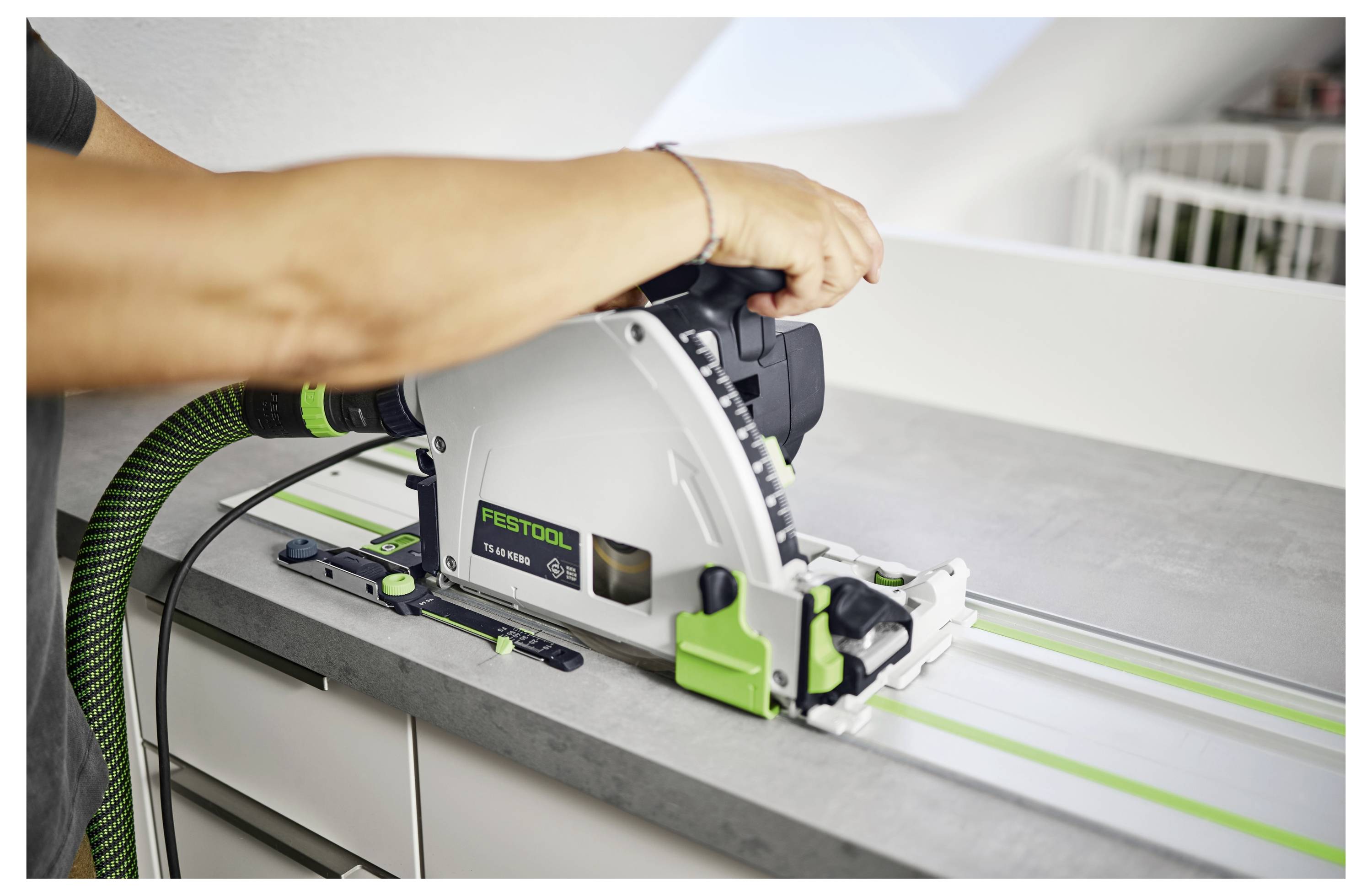 A person using a Festool circular saw on a countertop, guided by a green track for precise cutting. Bright indoor setting.