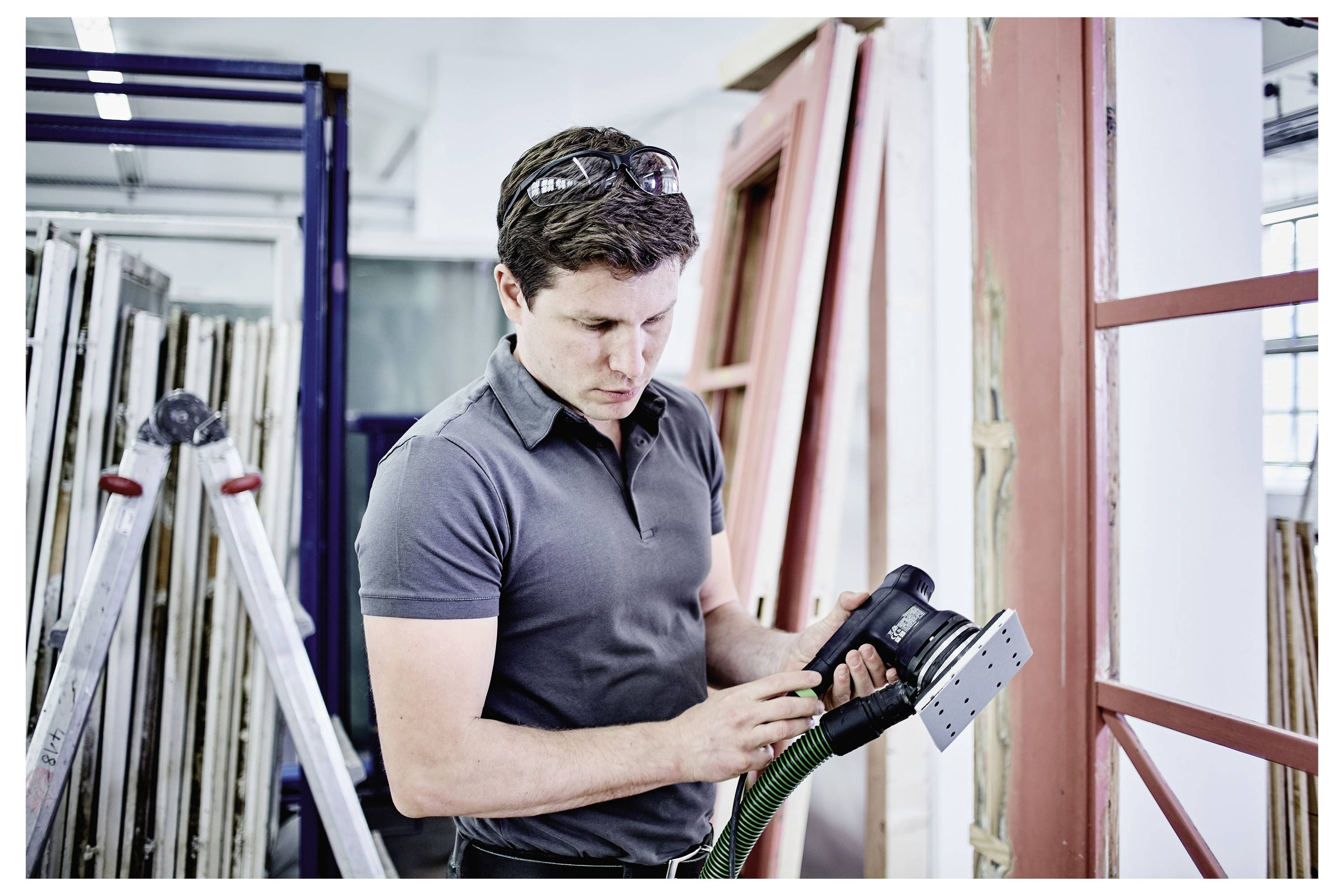 A person in a workshop uses a power sander on a window frame, surrounded by various building materials and tools.