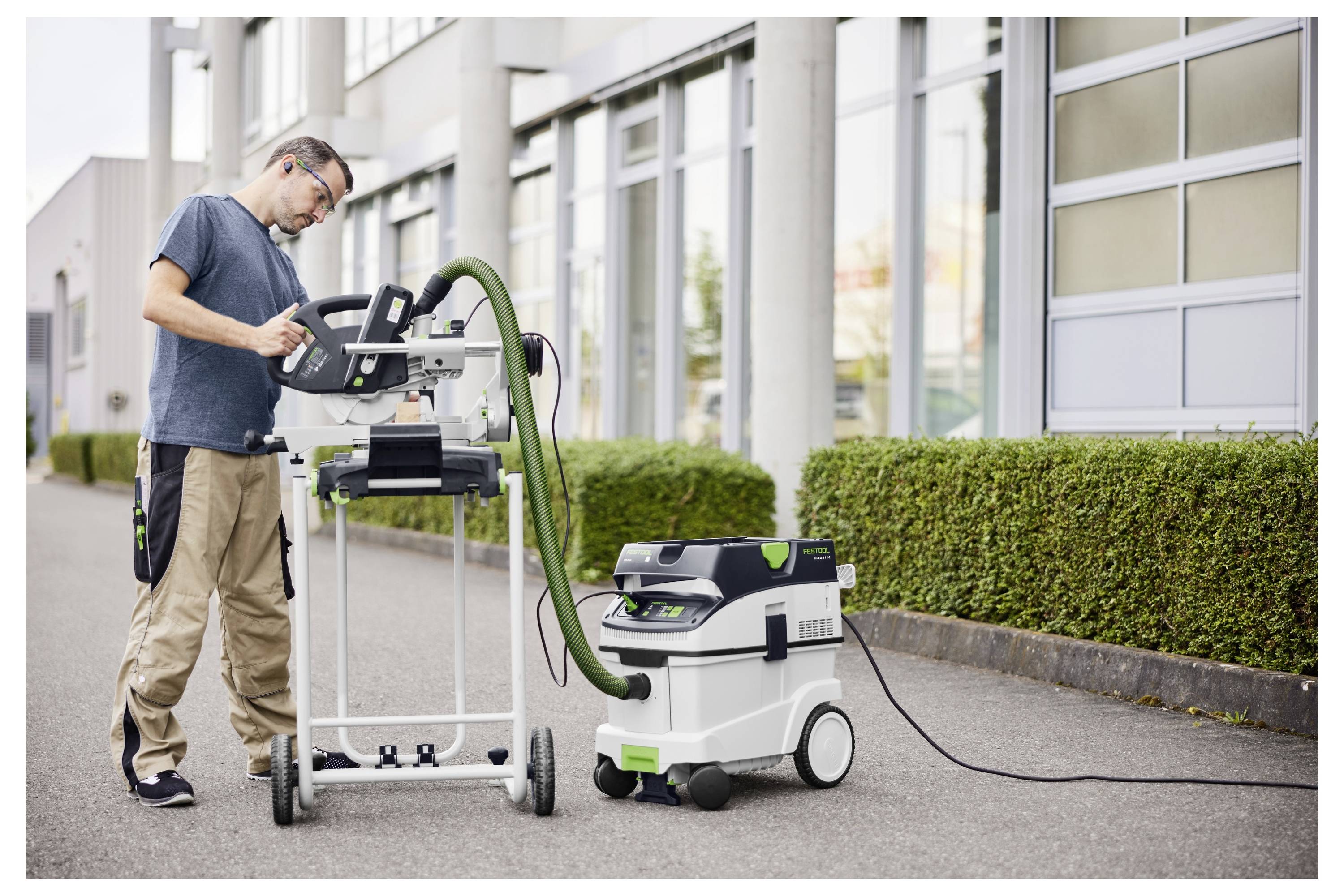 A man operates a miter saw connected to a dust extractor on a city sidewalk. He wears safety glasses and appears focused on his work.