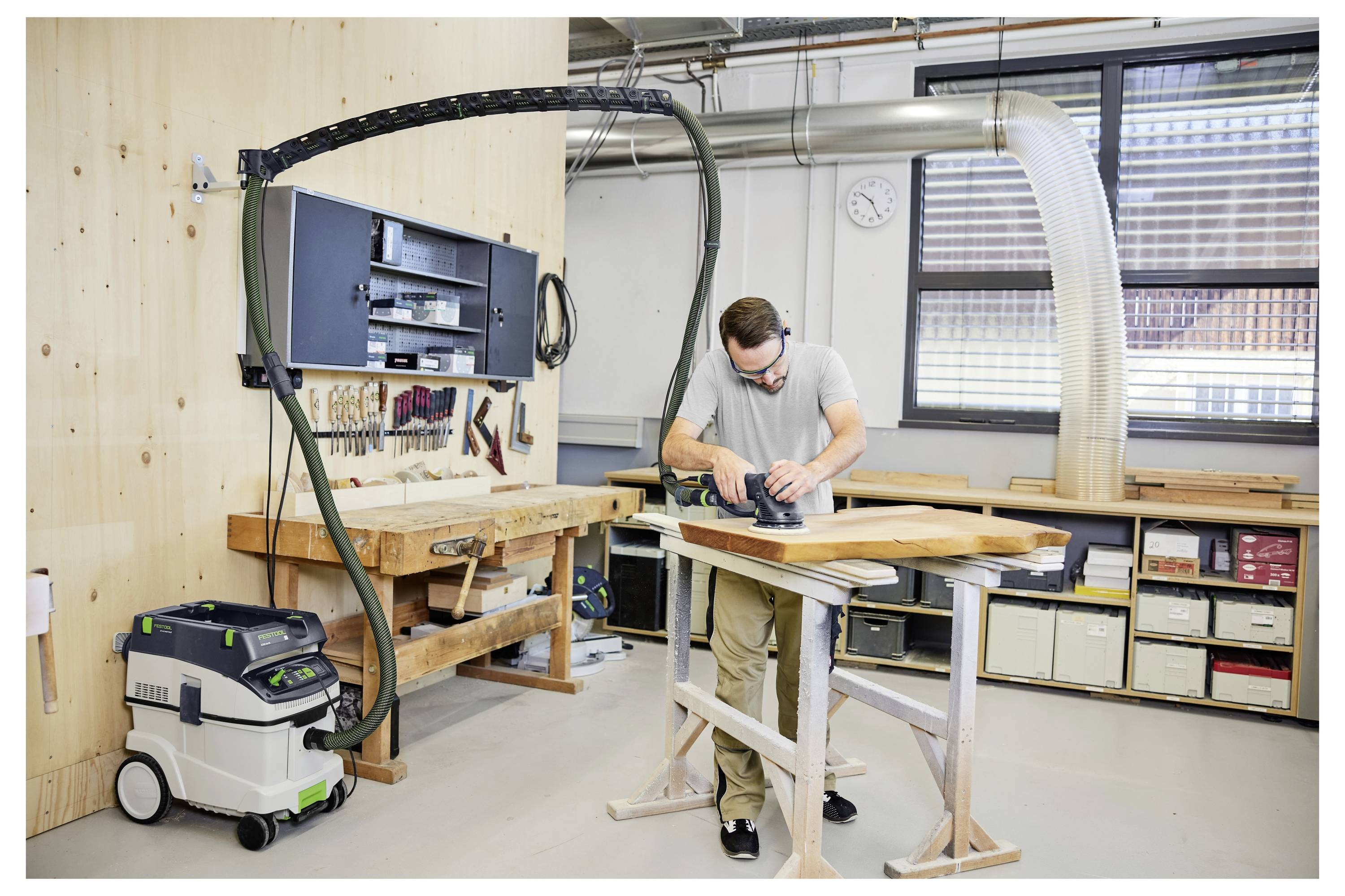A person using a power sander on a wooden board in a workshop equipped with tools, workbenches, and a dust extraction system.