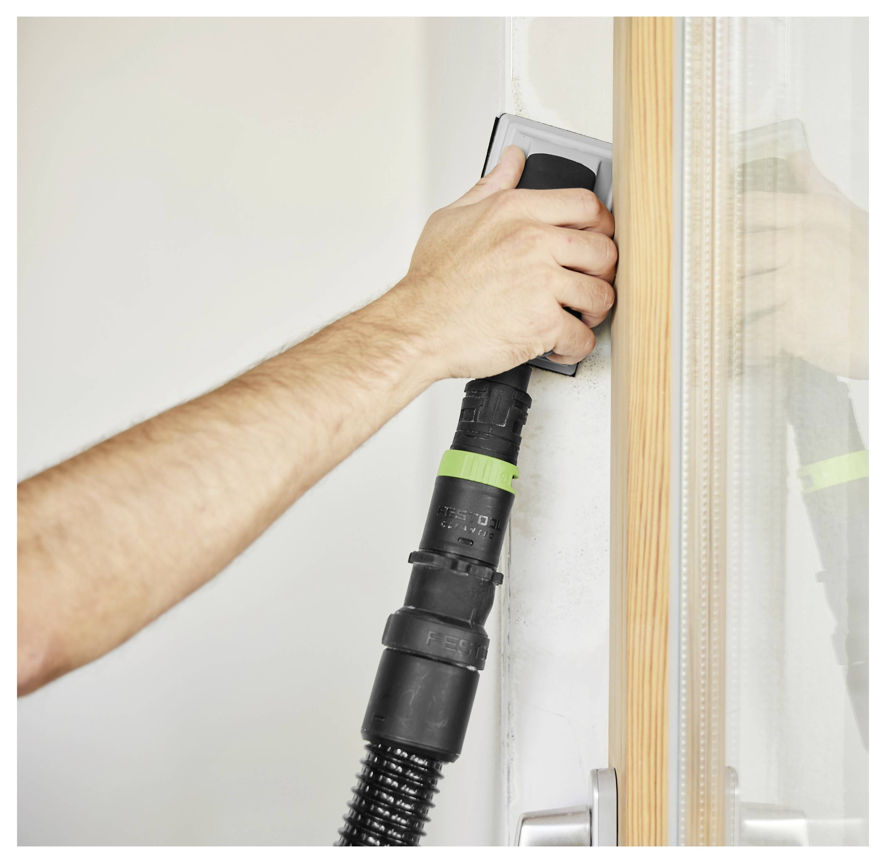 A person sanding a wall corner using a handheld electric sander, with a wooden doorframe and glass shower door partially visible.