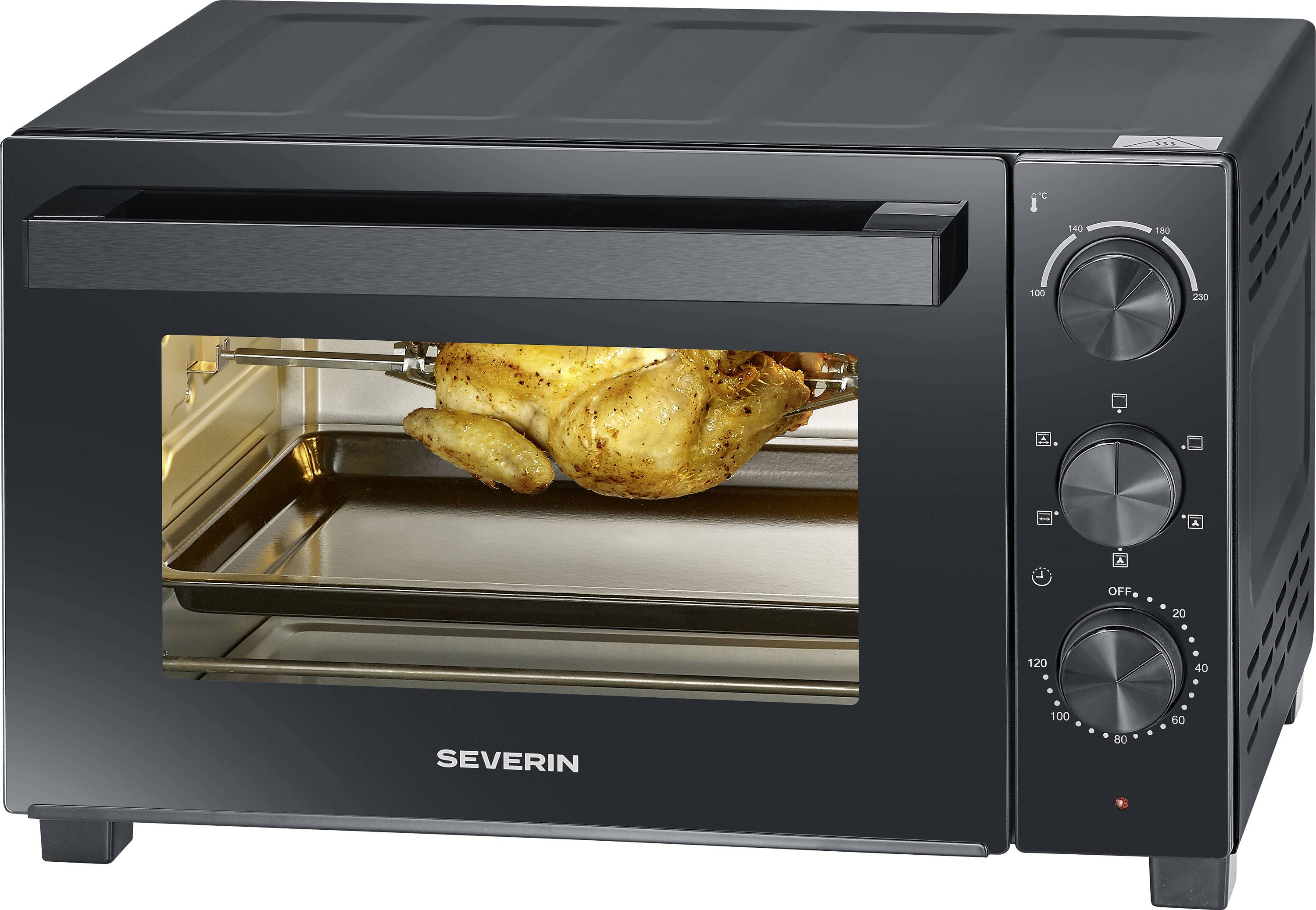A black countertop oven with a glass door, cooking a seasoned chicken on a rotisserie spit. Knobs on the side control temperature and settings.