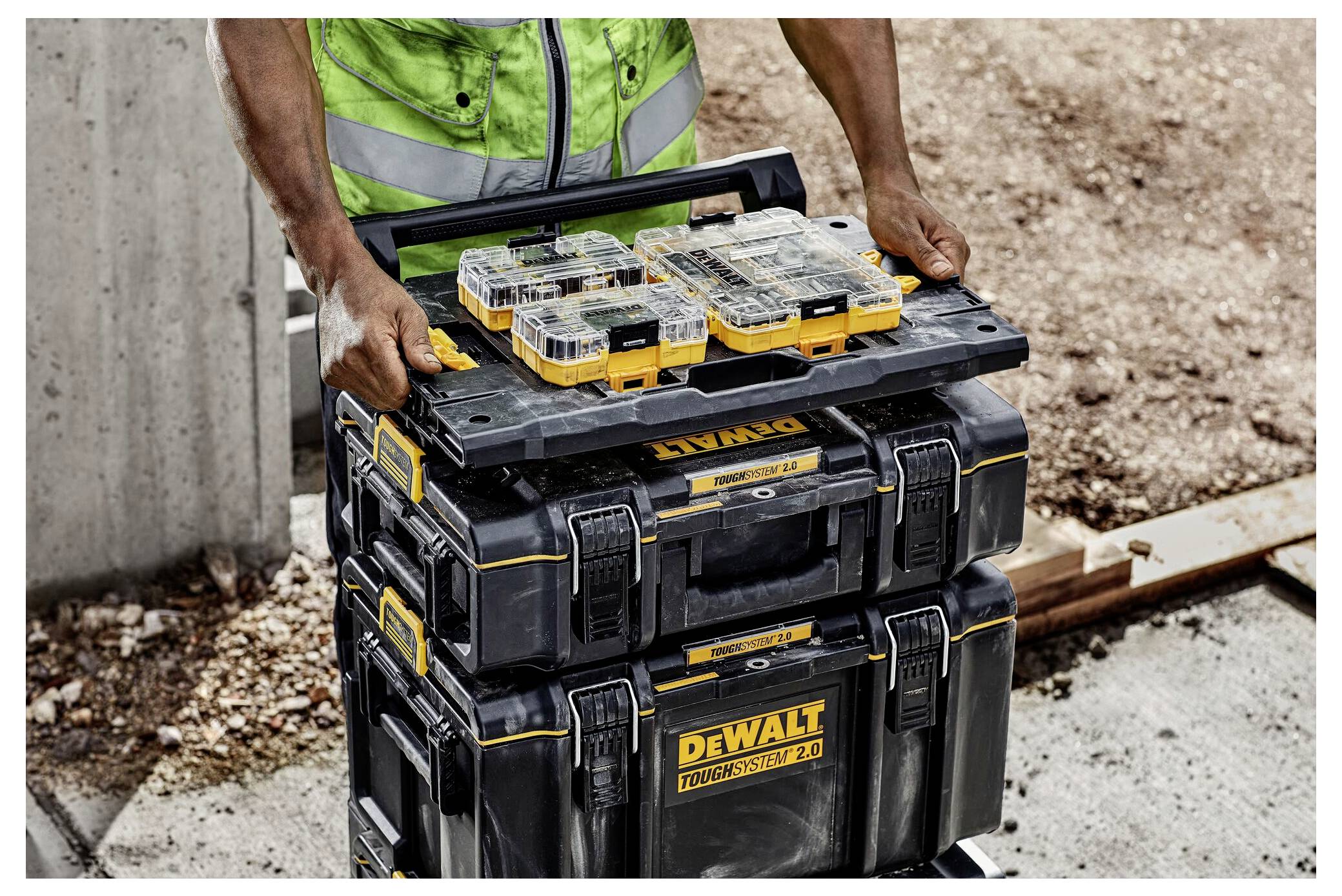 A worker in a green safety vest lifts a DeWalt ToughSystem 2.0 stack, showing several yellow and black toolboxes with tools inside.