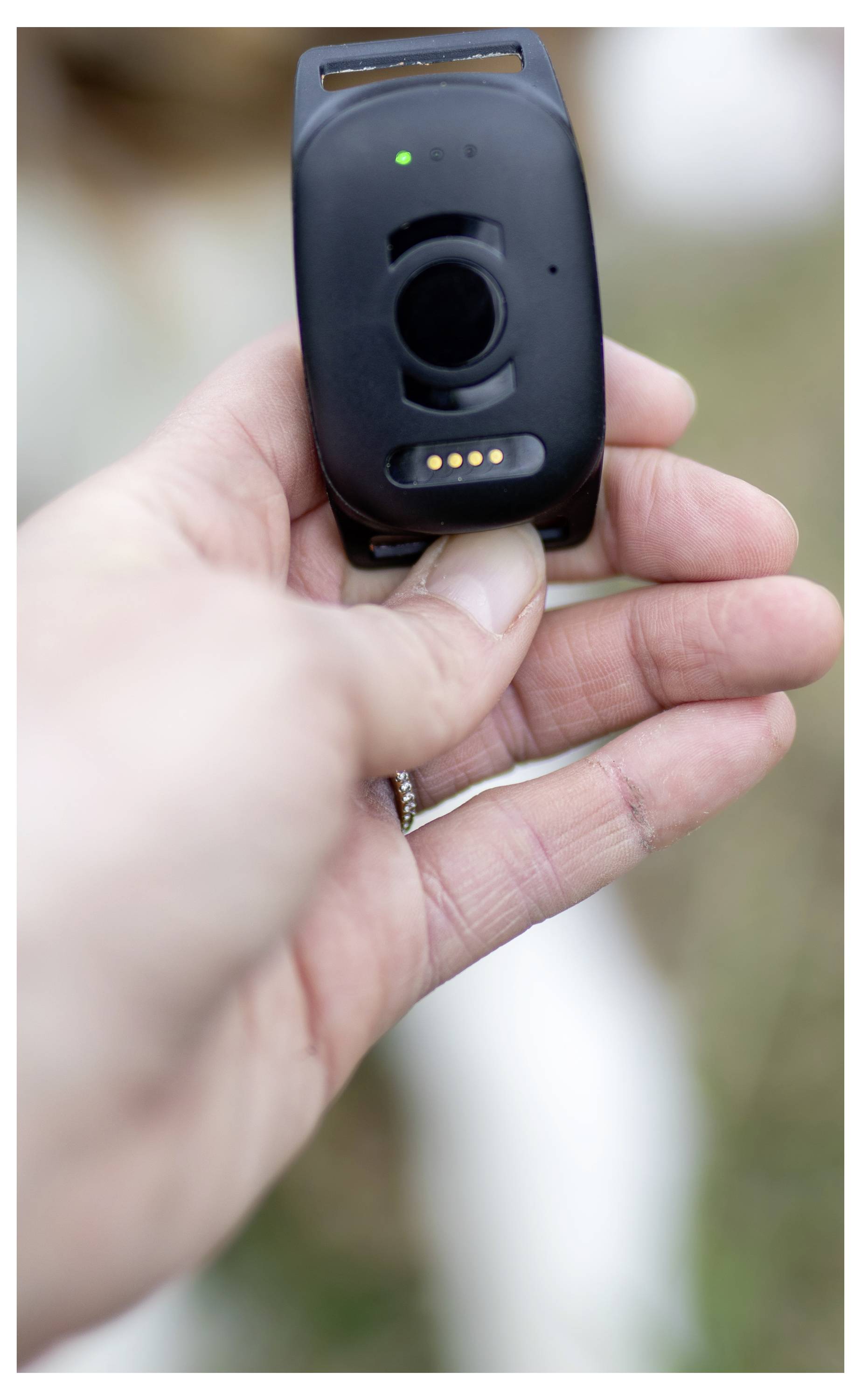 A hand holding a black GPS tracking device outdoors, possibly for a pet or person, with a green status light indicating it's active.