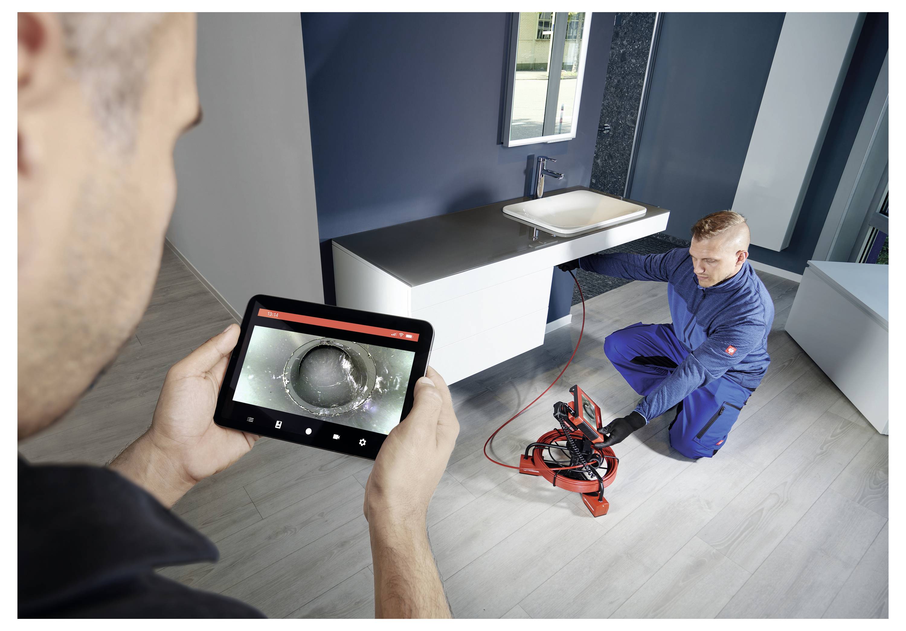 A plumber inspects a bathroom pipe using a camera and tablet display. Another person monitors the feed on the tablet screen.