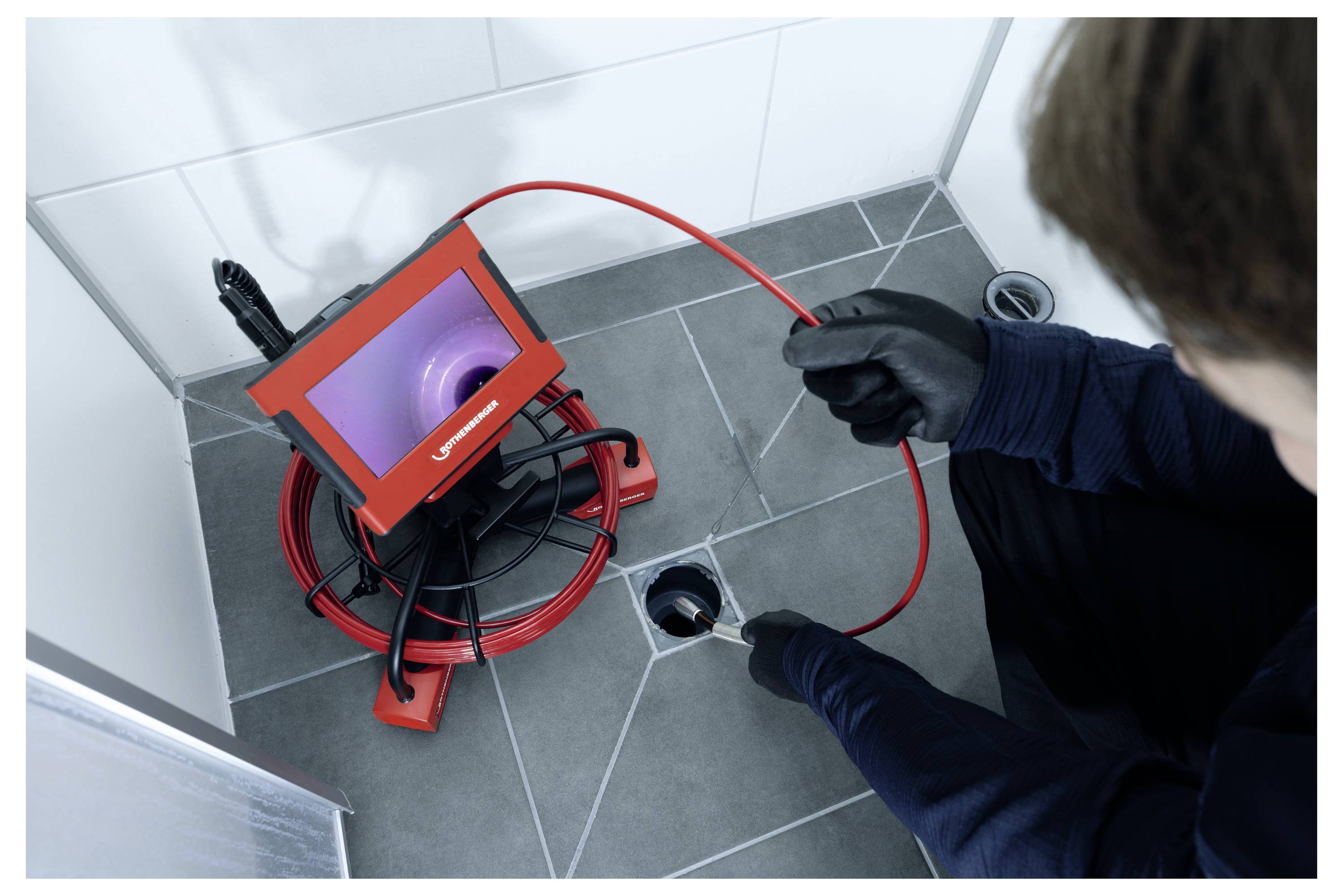 'A person uses a camera system to inspect a drainpipe on a tiled floor, viewing the pipe's interior on a display screen.'