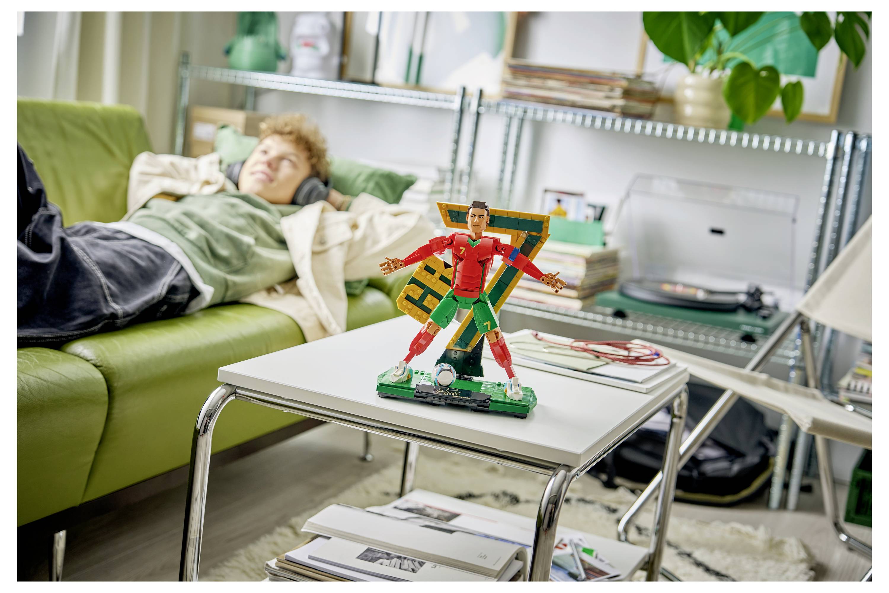 A person lies resting on a green couch while a colorful superhero toy figure stands on a coffee table in the foreground.