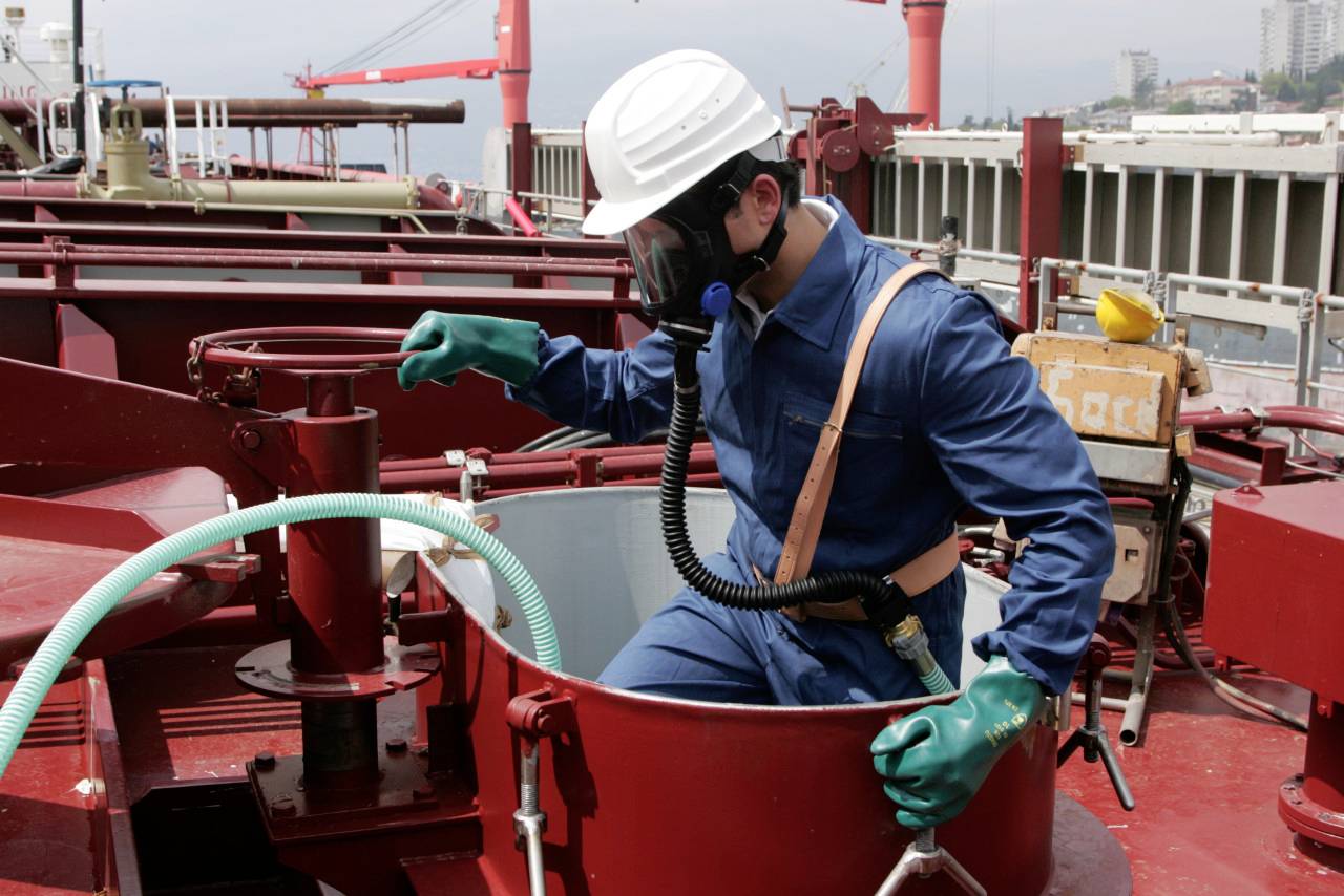 A worker in protective gear, including a helmet, gloves, and a respirator, climbs into a hatch on an industrial ship deck.