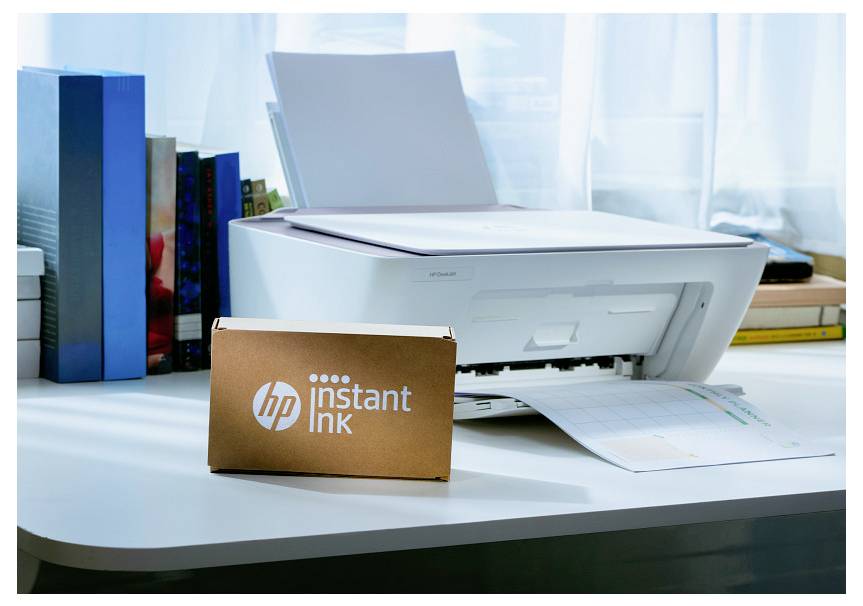 A white printer on a desk, next to books, with an HP Instant Ink box in the foreground. Light filters through a window behind.