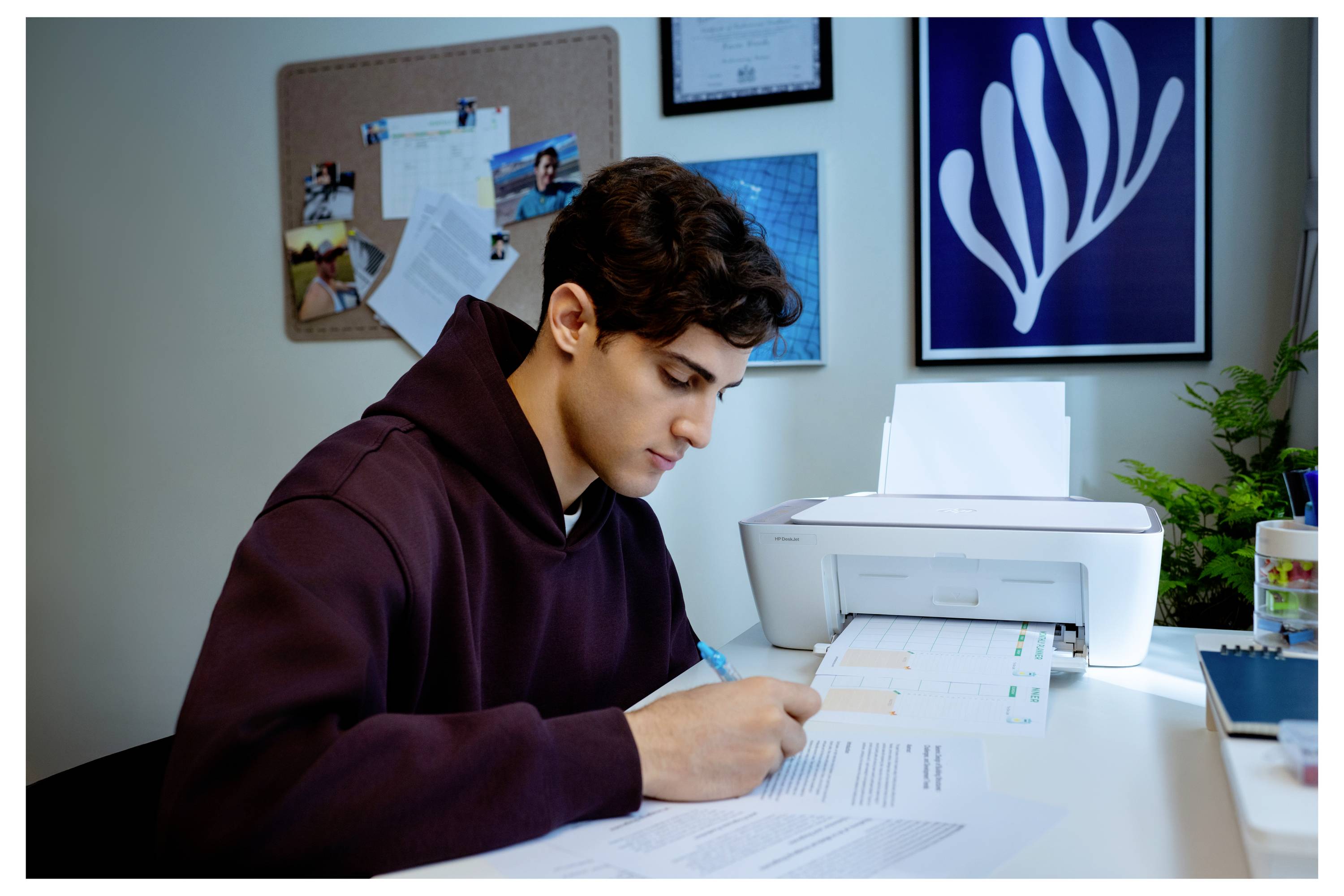 A person in a hoodie sits at a desk, writing on papers with a printer nearby. The wall displays a bulletin board and framed artwork.