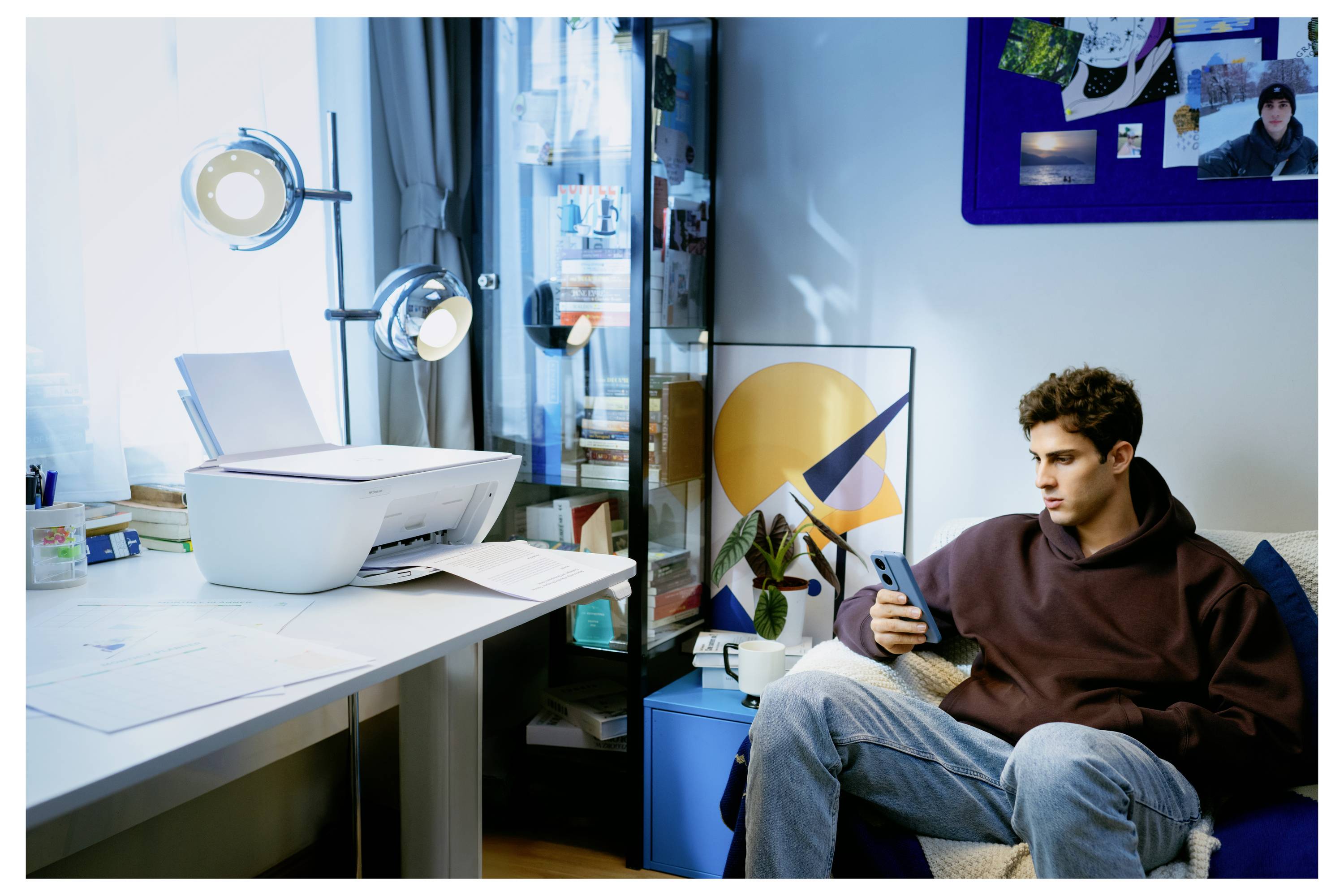 A person sits on a couch looking at a smartphone, beside a table with a printer and papers. A cabinet with books and art decor is nearby.