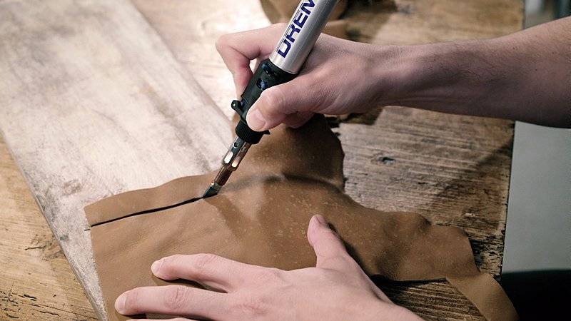 A person uses a soldering iron to cut through brown leather on a wooden table.