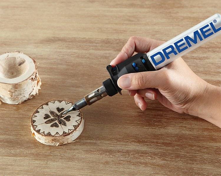 A hand using a Dremel tool to etch a floral design on a wooden disc, with another blank disc nearby on a wooden surface.