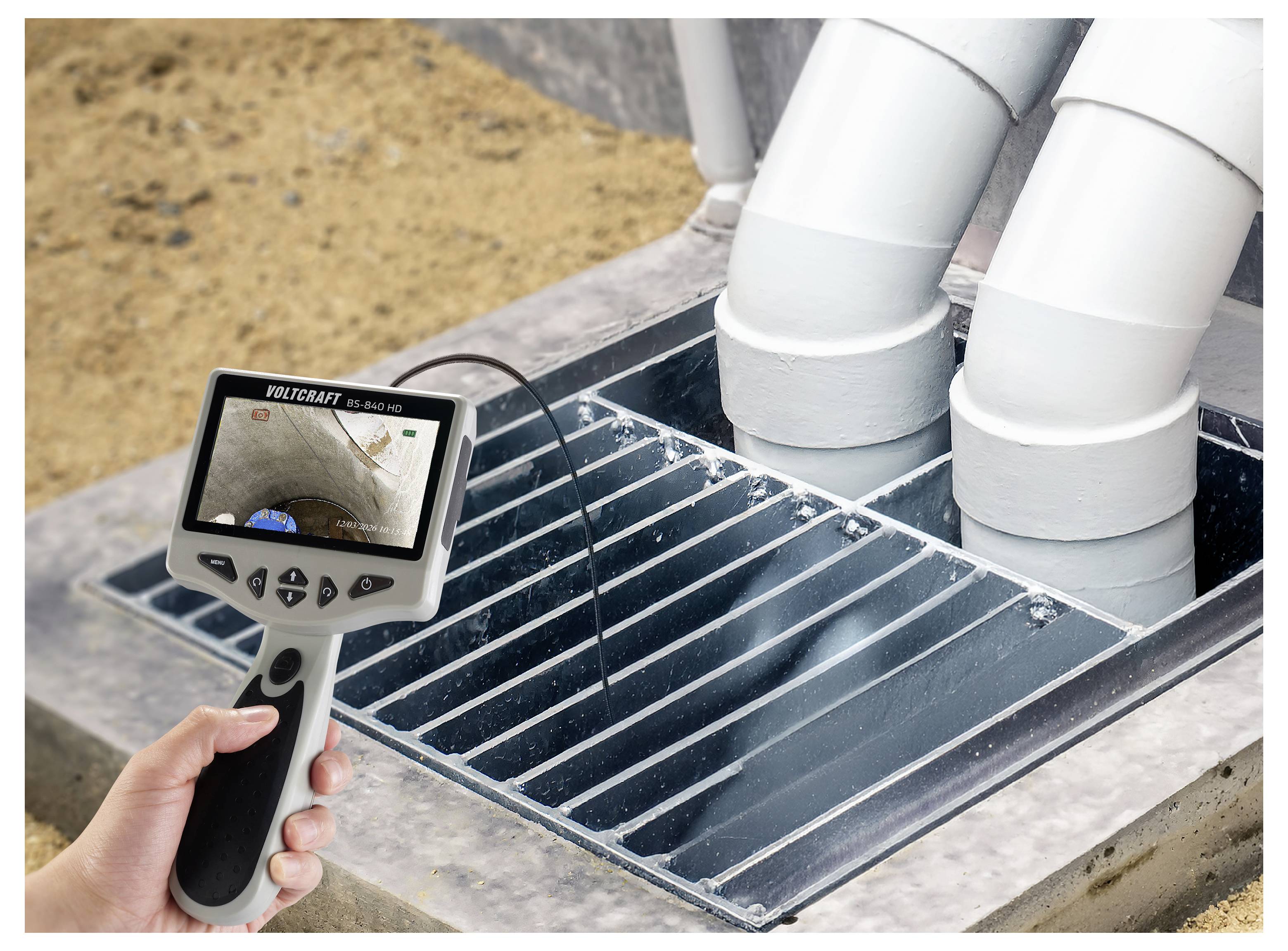 Handheld inspection camera shows inside a drain opening with white pipes, used to inspect underground plumbing or sewer lines.