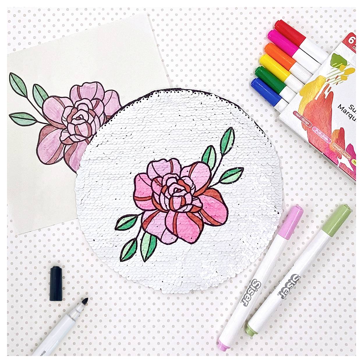 A completed sequin art project depicting a pink rose with green leaves, alongside markers and a reference drawing.