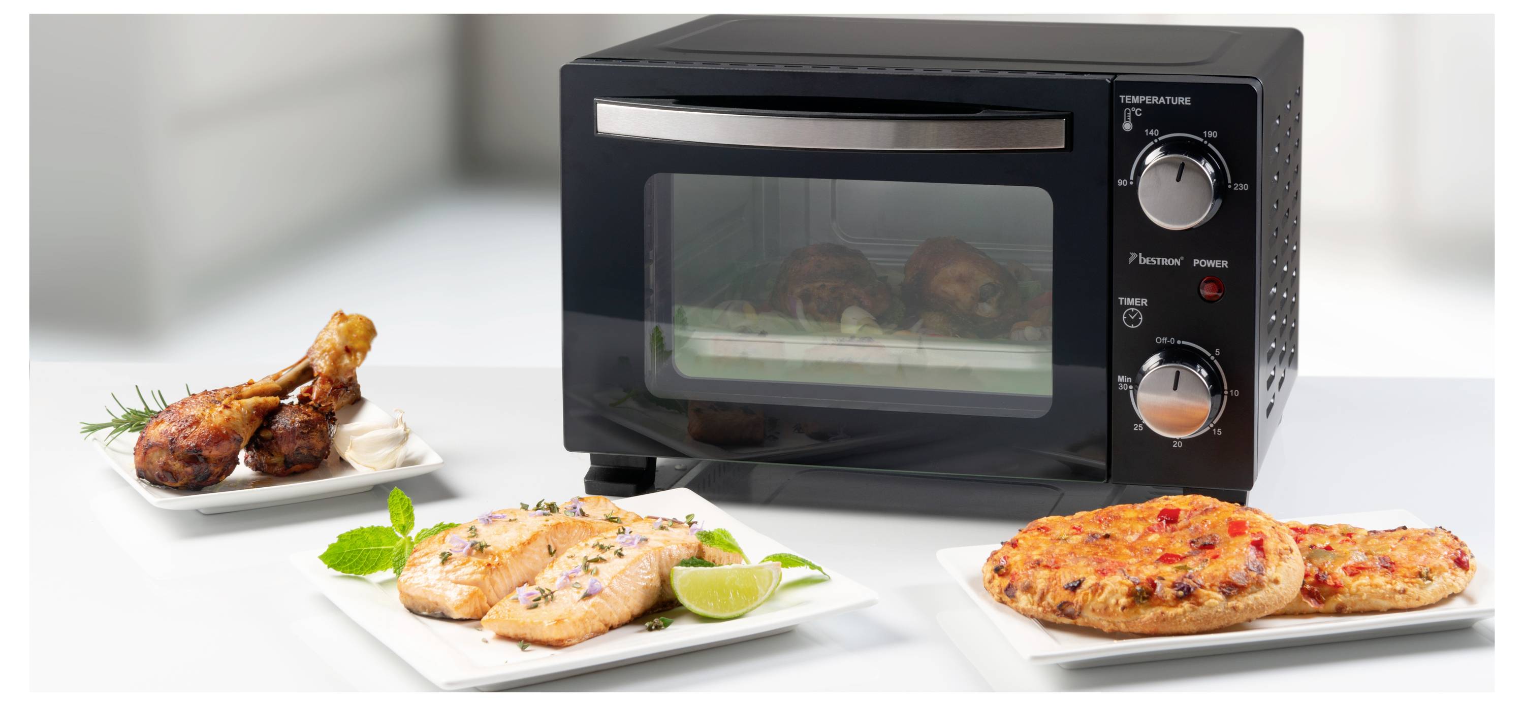 A black countertop oven with two control knobs is surrounded by plates of prepared food: grilled chicken, a small pizza, and lime-garnished fillets.