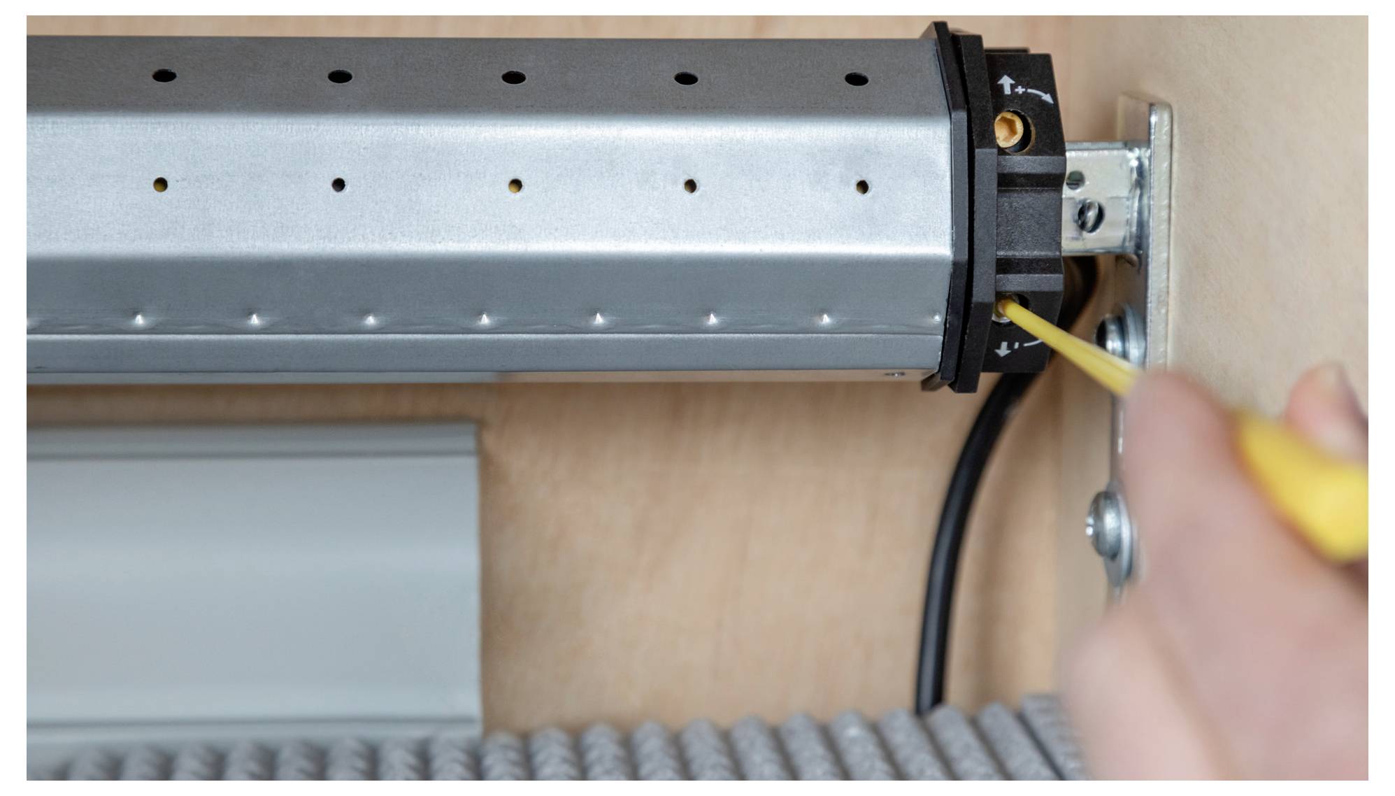 A person using a screwdriver on a metallic roller mechanism. The roller is partially enclosed by a wooden casing and a floor mat is visible below.