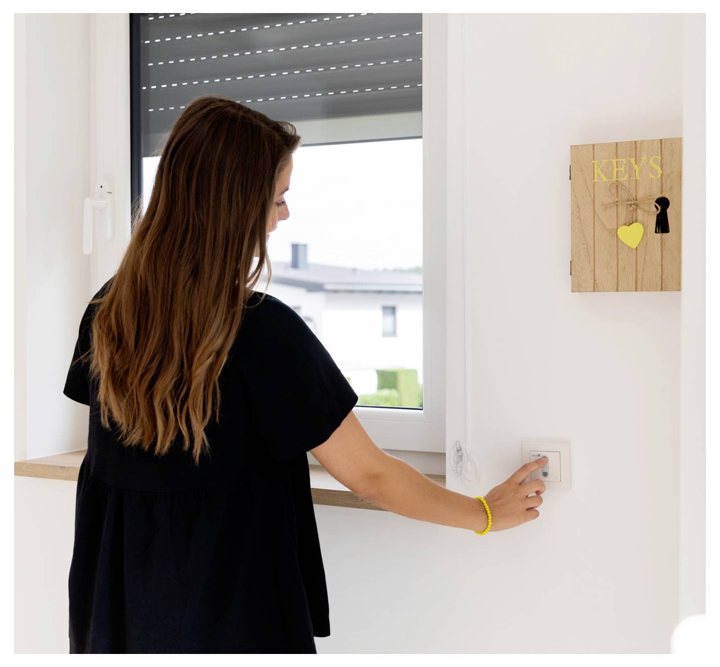 A woman in a black shirt presses a button on a wall switch under a window. A wooden key holder labeled 'KEYS' with a keyhole is nearby.
