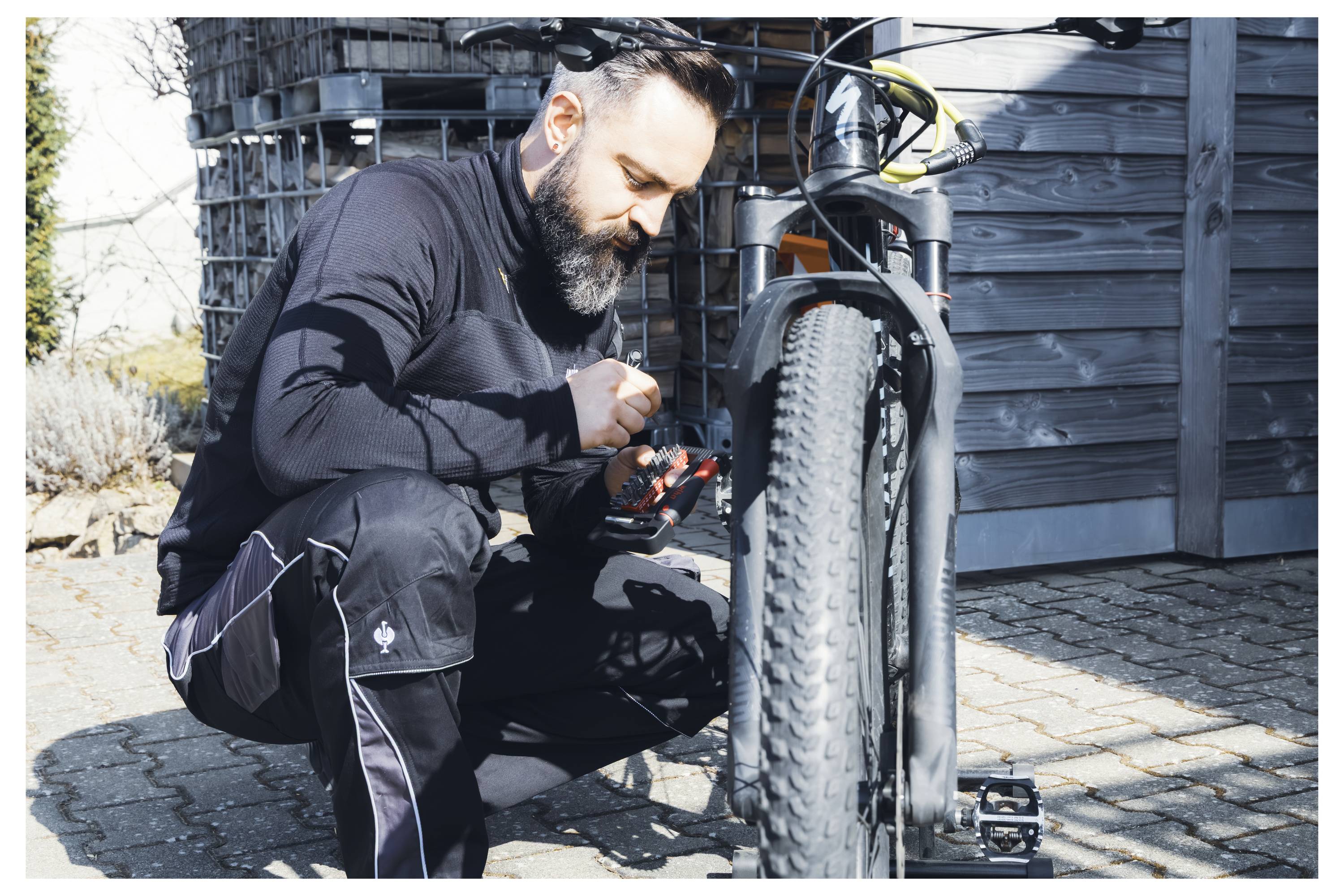 A man kneels beside a bicycle, adjusting gears with a tool. He's wearing black clothing, focusing intently, outdoors on a sunny day.