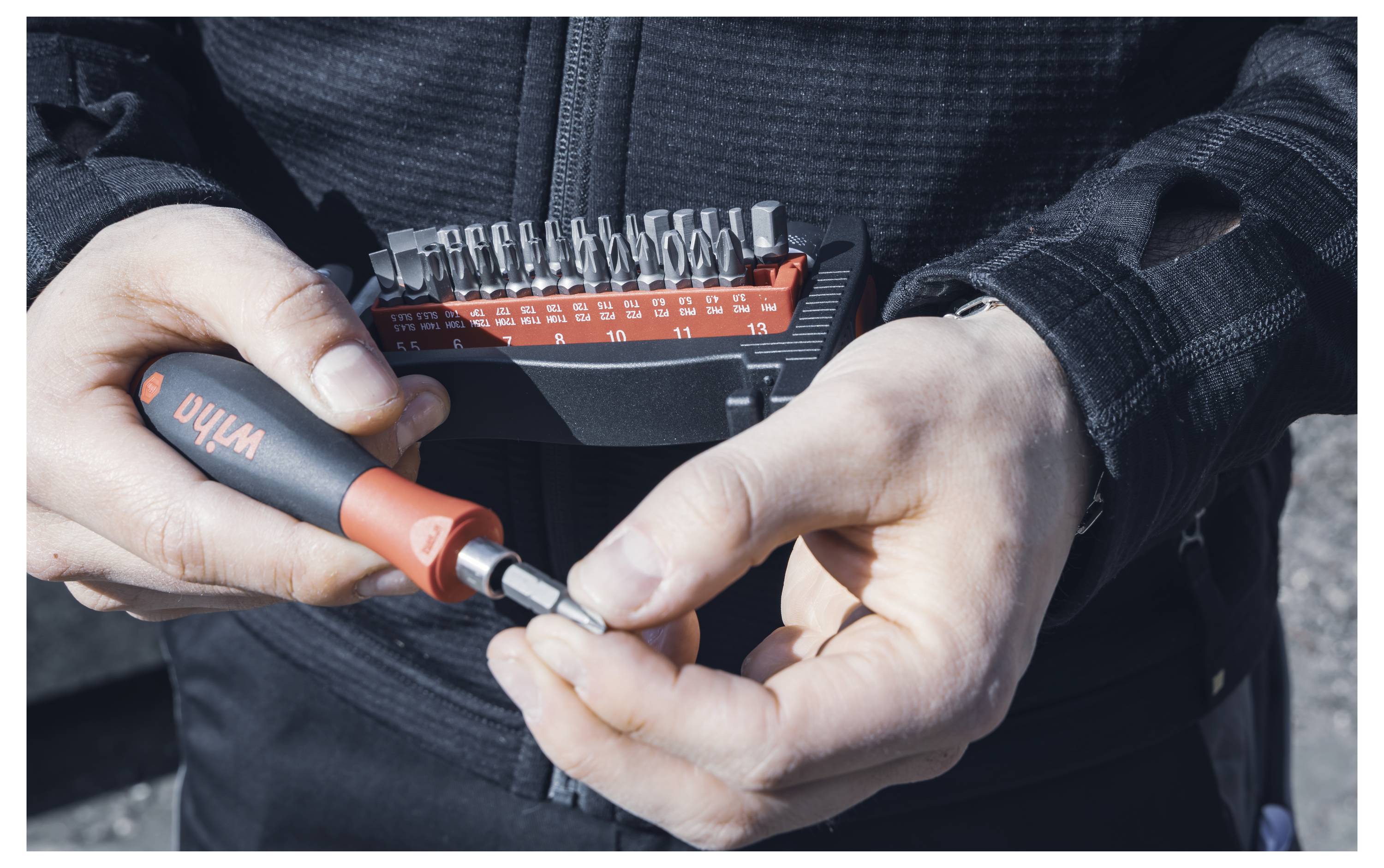 A person wearing dark clothing holds a red and black tool set with interchangeable bits, preparing to attach a bit to the handle.