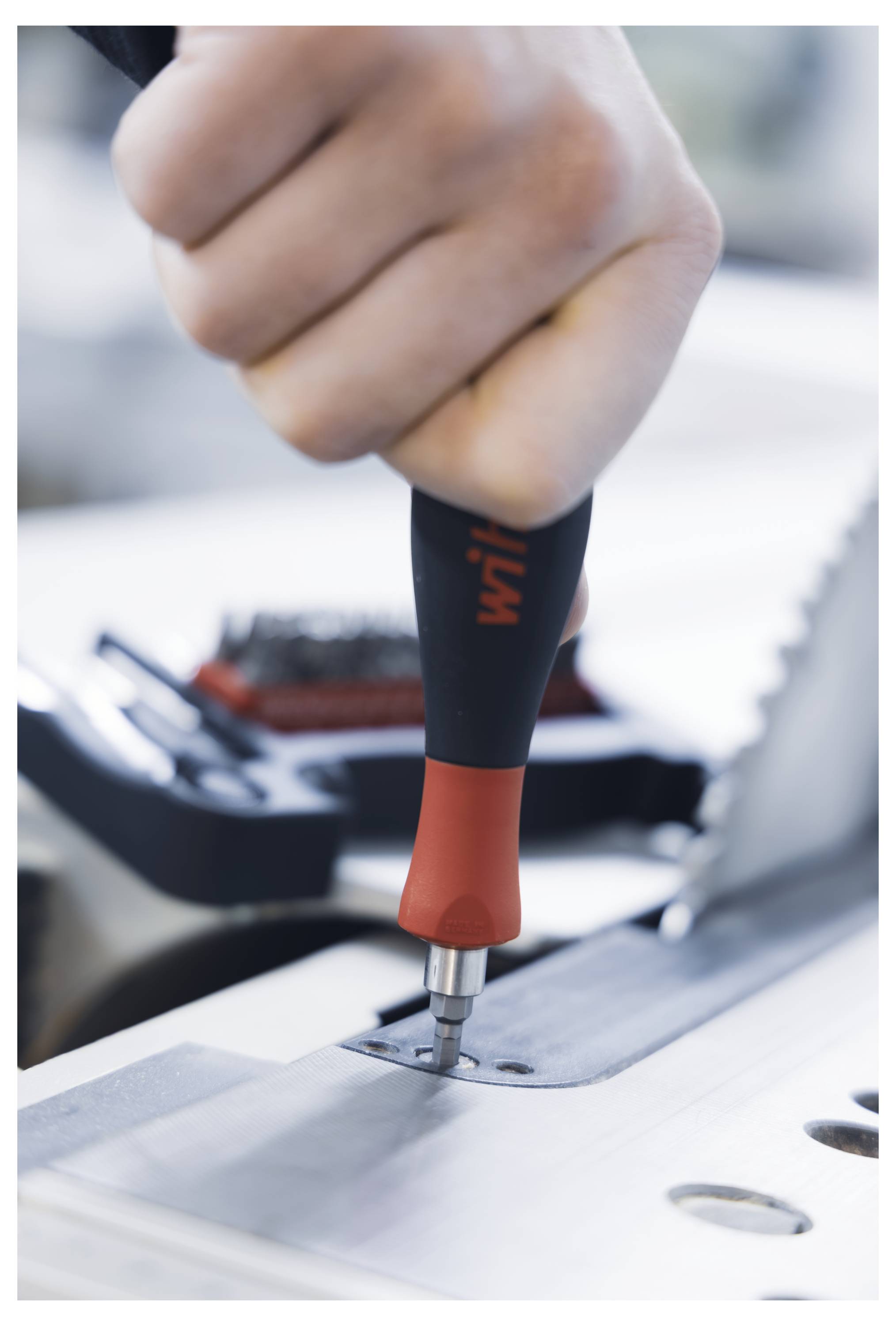 A hand using a red and black screwdriver to tighten a screw on a metal surface, with a blurred saw blade in the background.