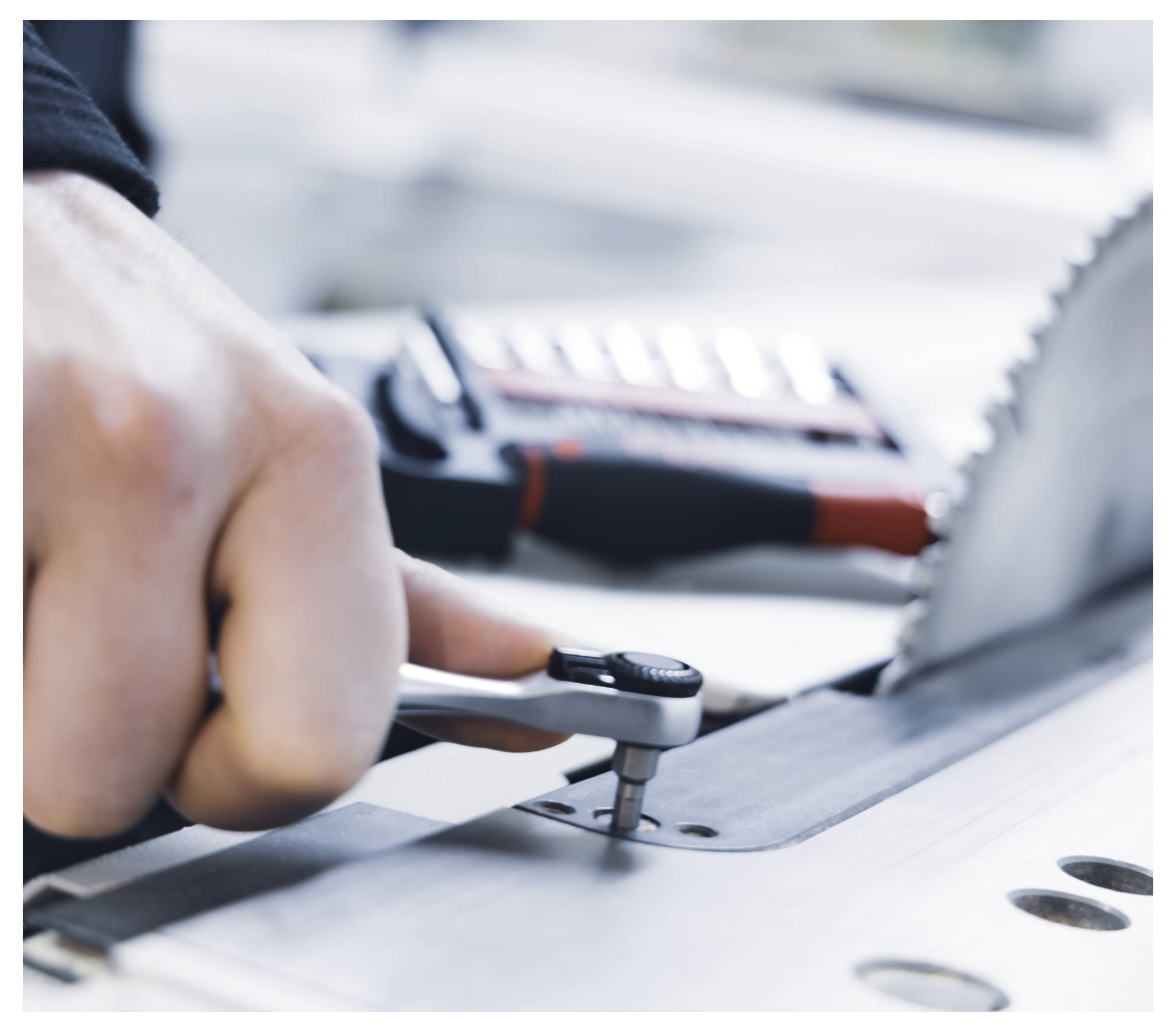 A hand uses a wrench to adjust a bolt near a circular saw blade, with a blurred toolbox in the background.